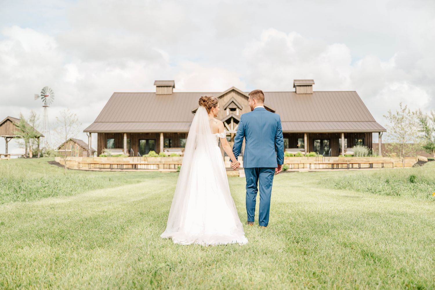 Bride and groom hold hands facing a rustic barn wedding venue on green lawn. The Old Barns at Dry Run Farms in Williamsport, Ohio.