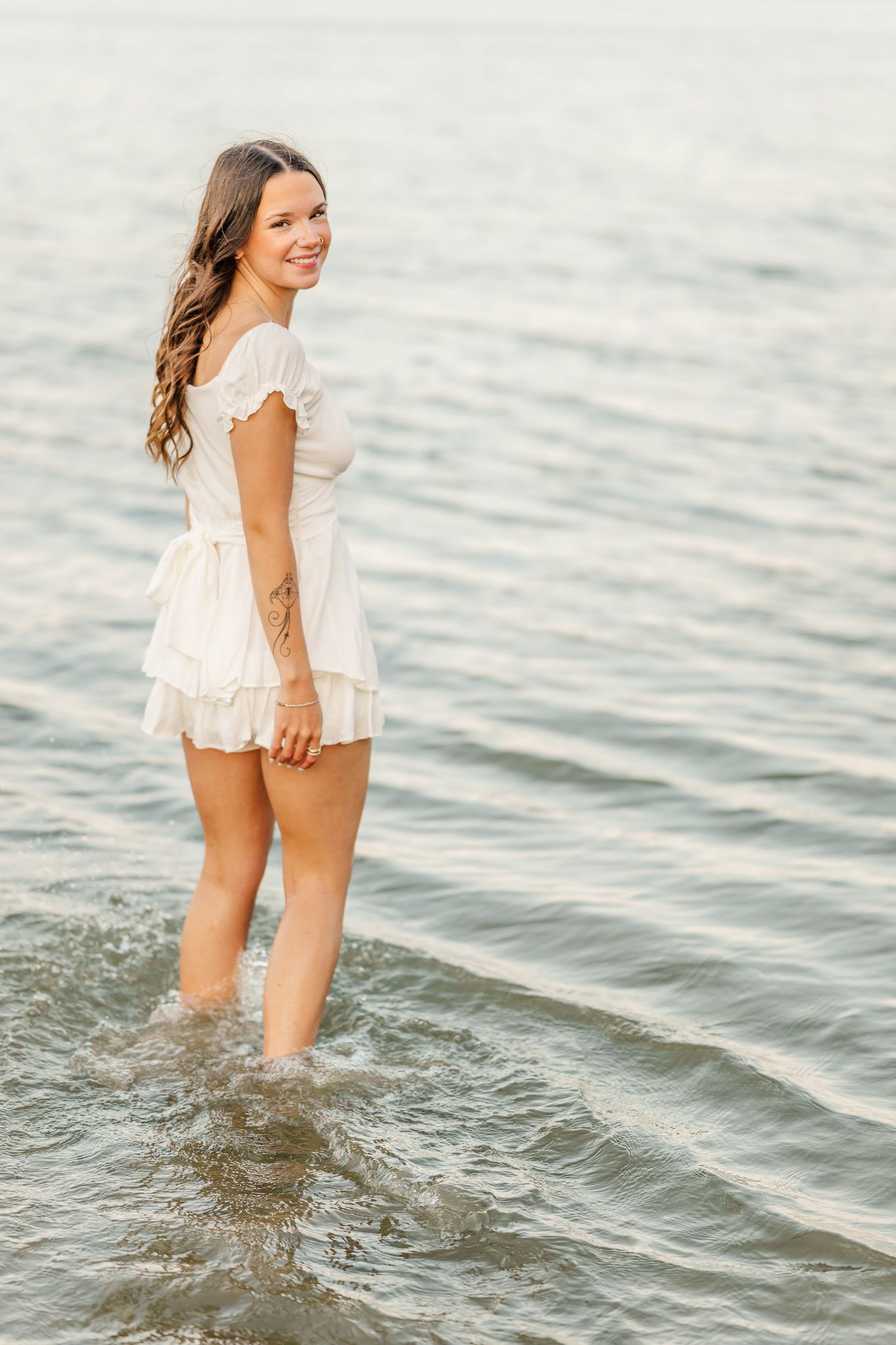Young woman in white ruffle dress standing in shallow ocean water, smiling over her shoulder at sunset. Alum Creek State Beach in Ohio.