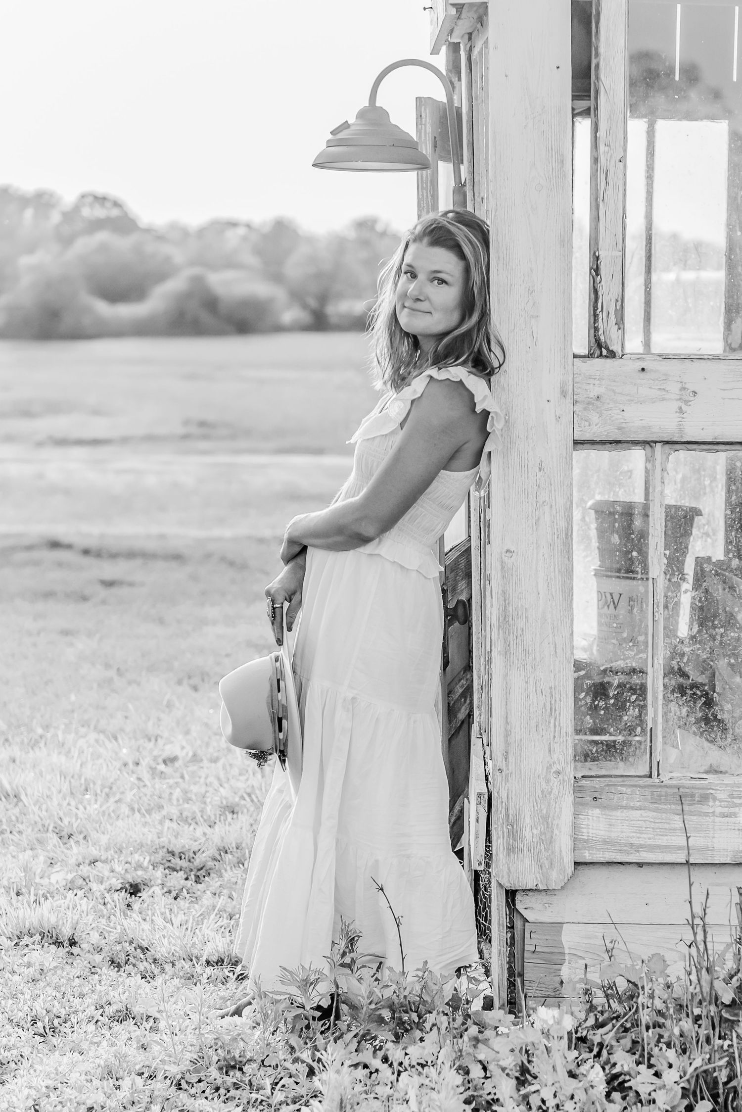 Black and white portrait of someone in a flowing dress leaning against a rustic wooden post with a lamp overhead.