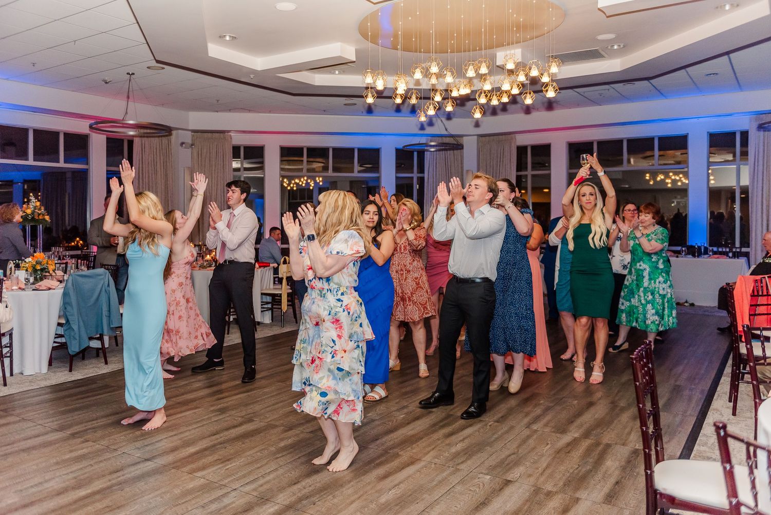 Wedding guests dancing together on a wooden dance floor under elegant chandeliers at a reception.