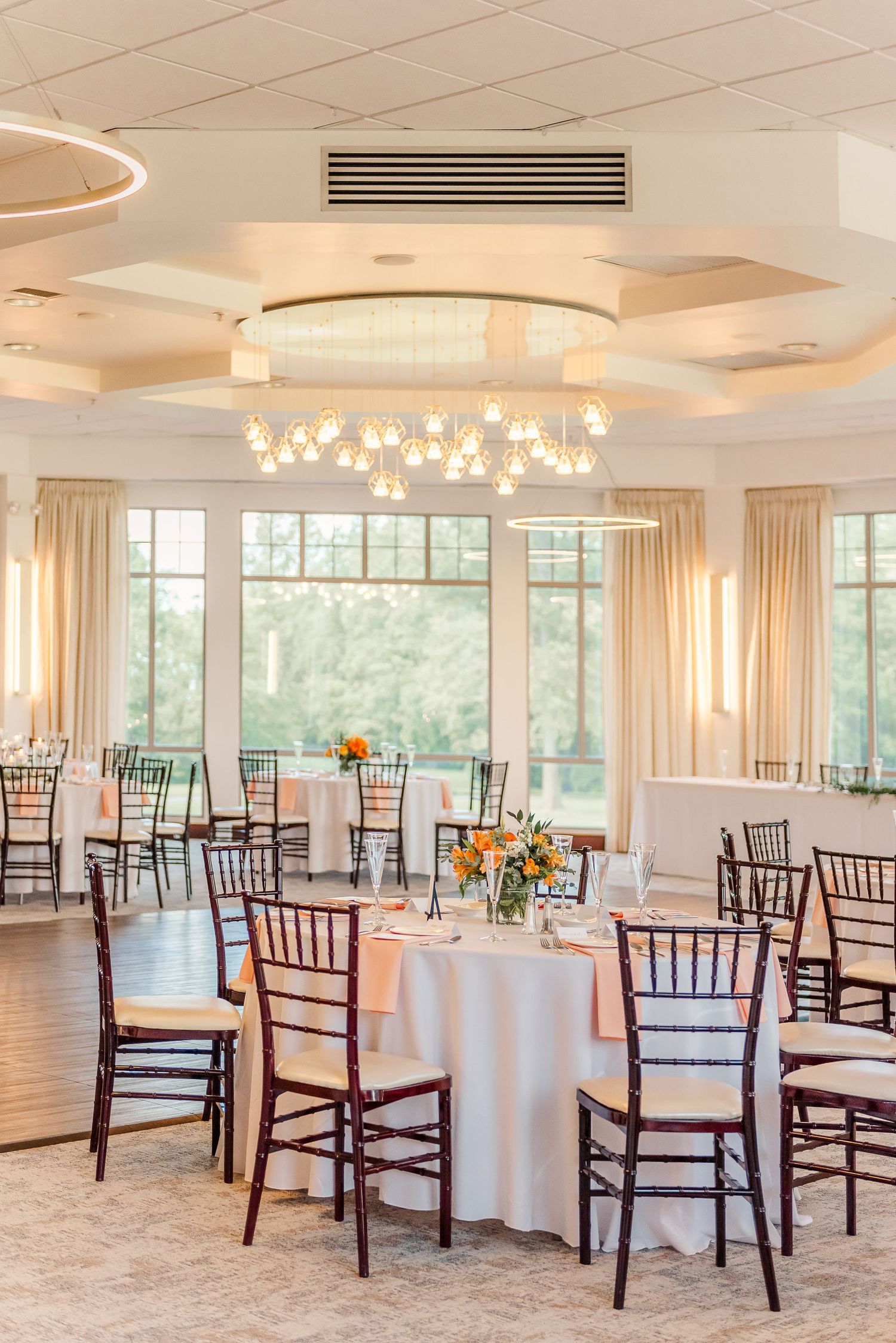 Elegant banquet hall setup with round tables, white linens, mahogany Chiavari chairs and natural light from large windows.