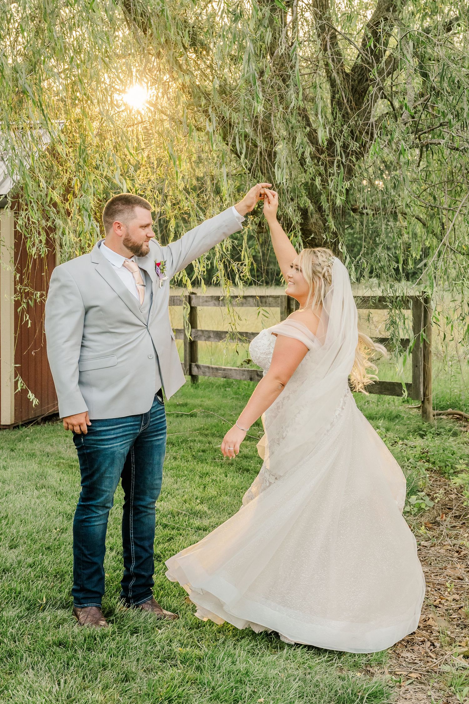 A couple dances and twirls under willow trees during golden hour in a rustic outdoor setting.
