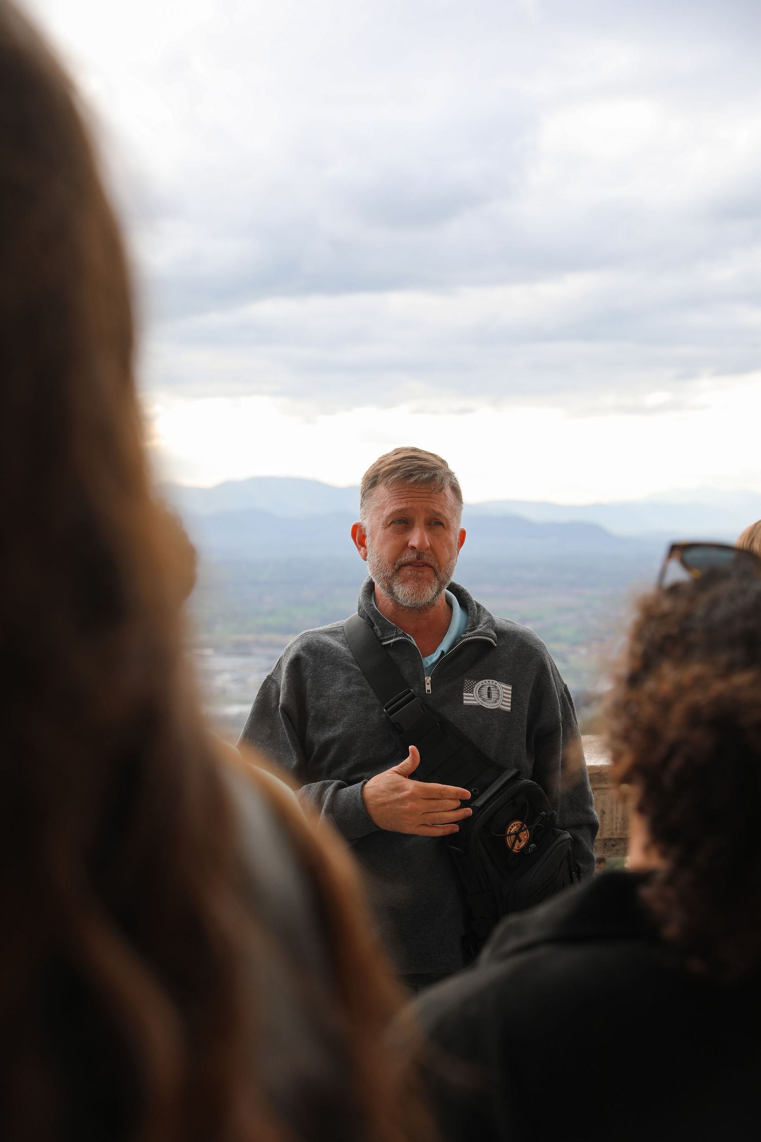 Man with gray beard speaks to group outdoors with mountain landscape backdrop under cloudy sky.