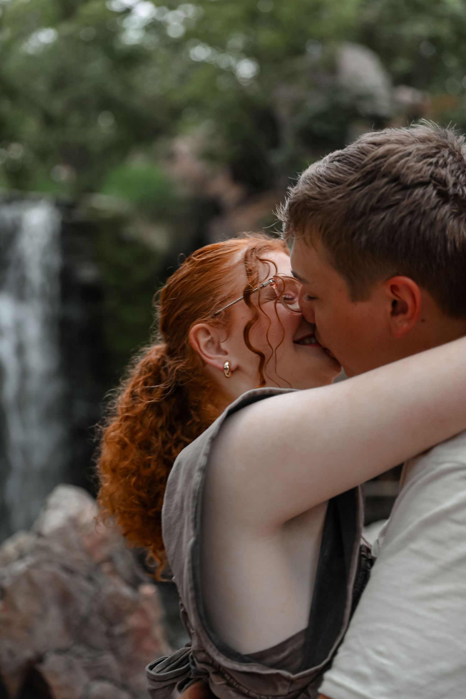 A couple shares a romantic kiss near a waterfall, the woman with red hair embracing a man outdoors.
