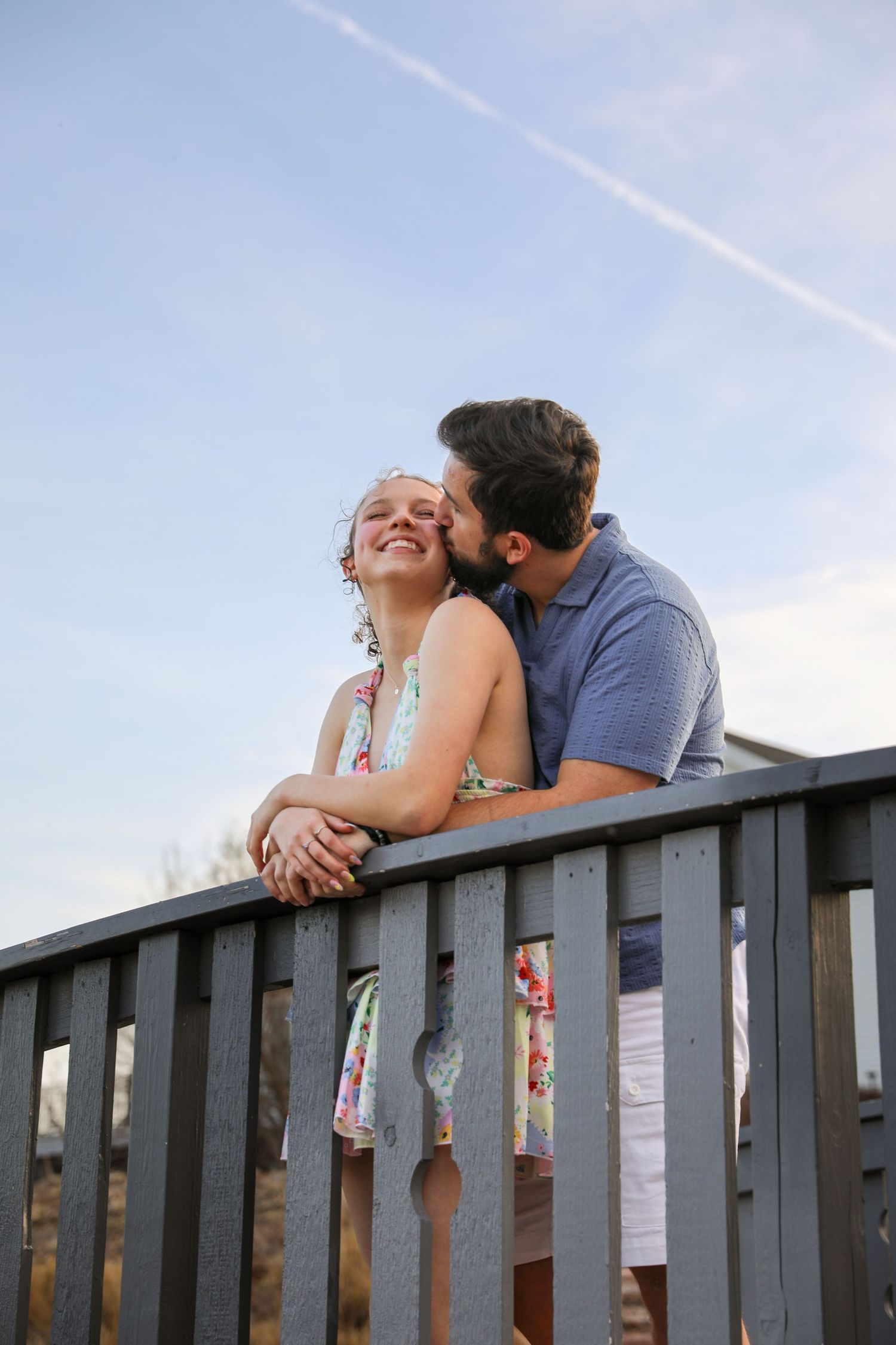 A couple shares an intimate moment while leaning over a wooden railing against a blue sky backdrop.