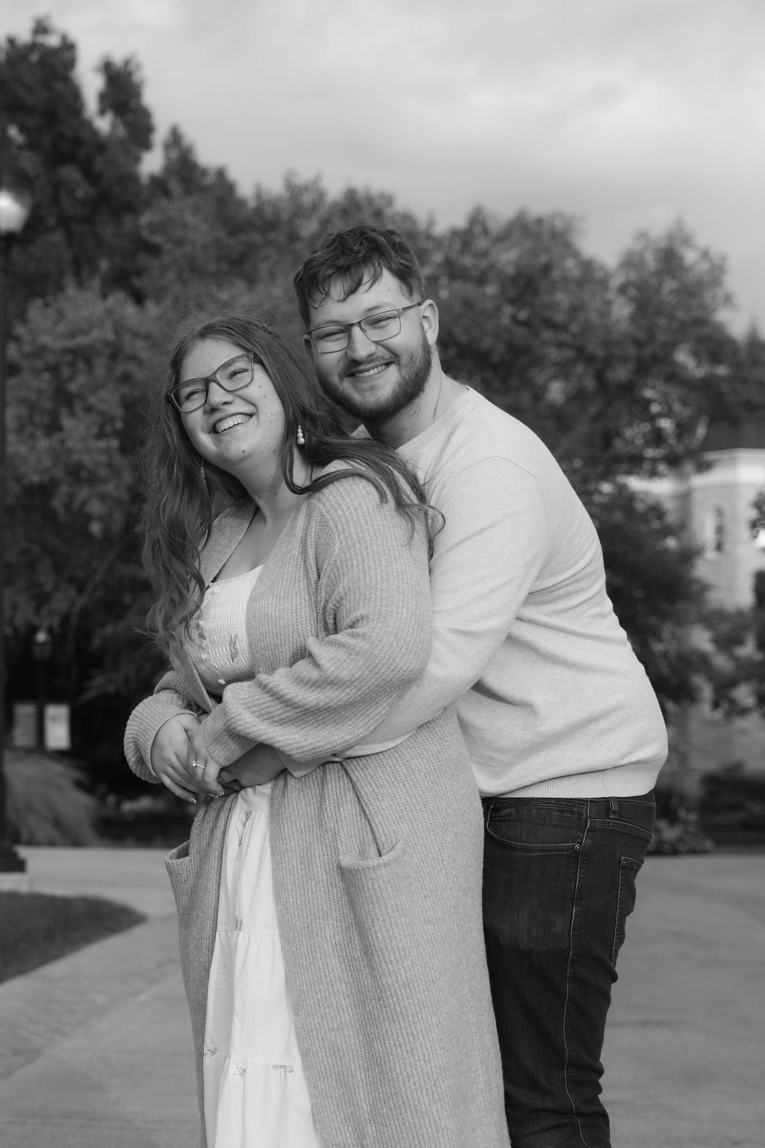 Black and white photo of a smiling couple embracing outdoors, woman in cardigan, man in sweater, both wearing glasses.