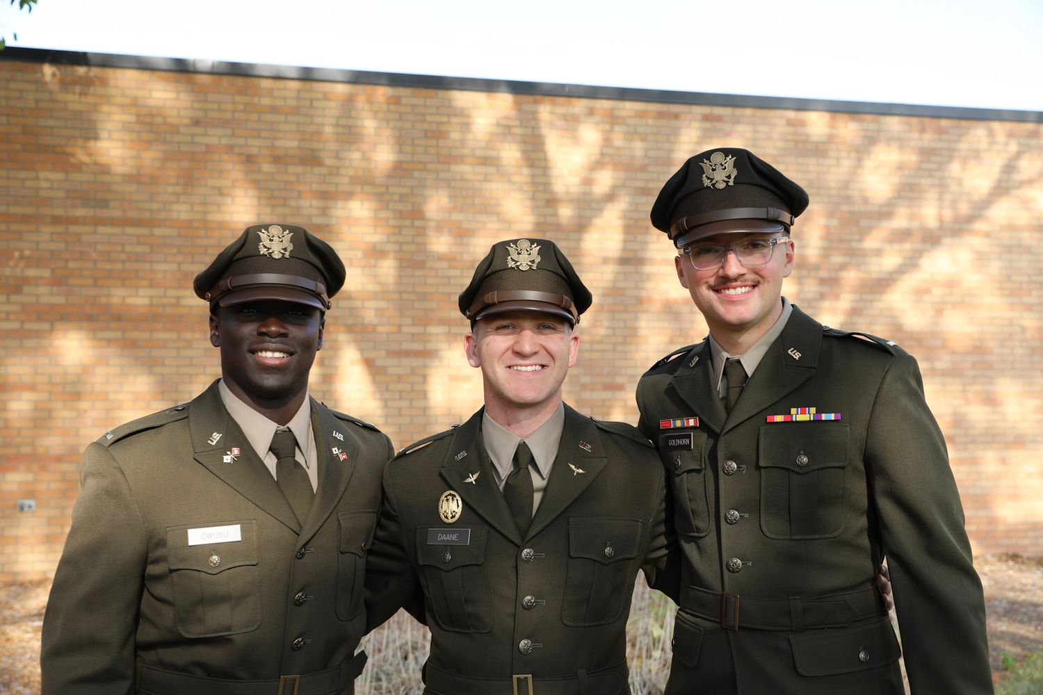 Three men in olive green U.S. Army dress uniforms smile together outdoors near a brick wall.