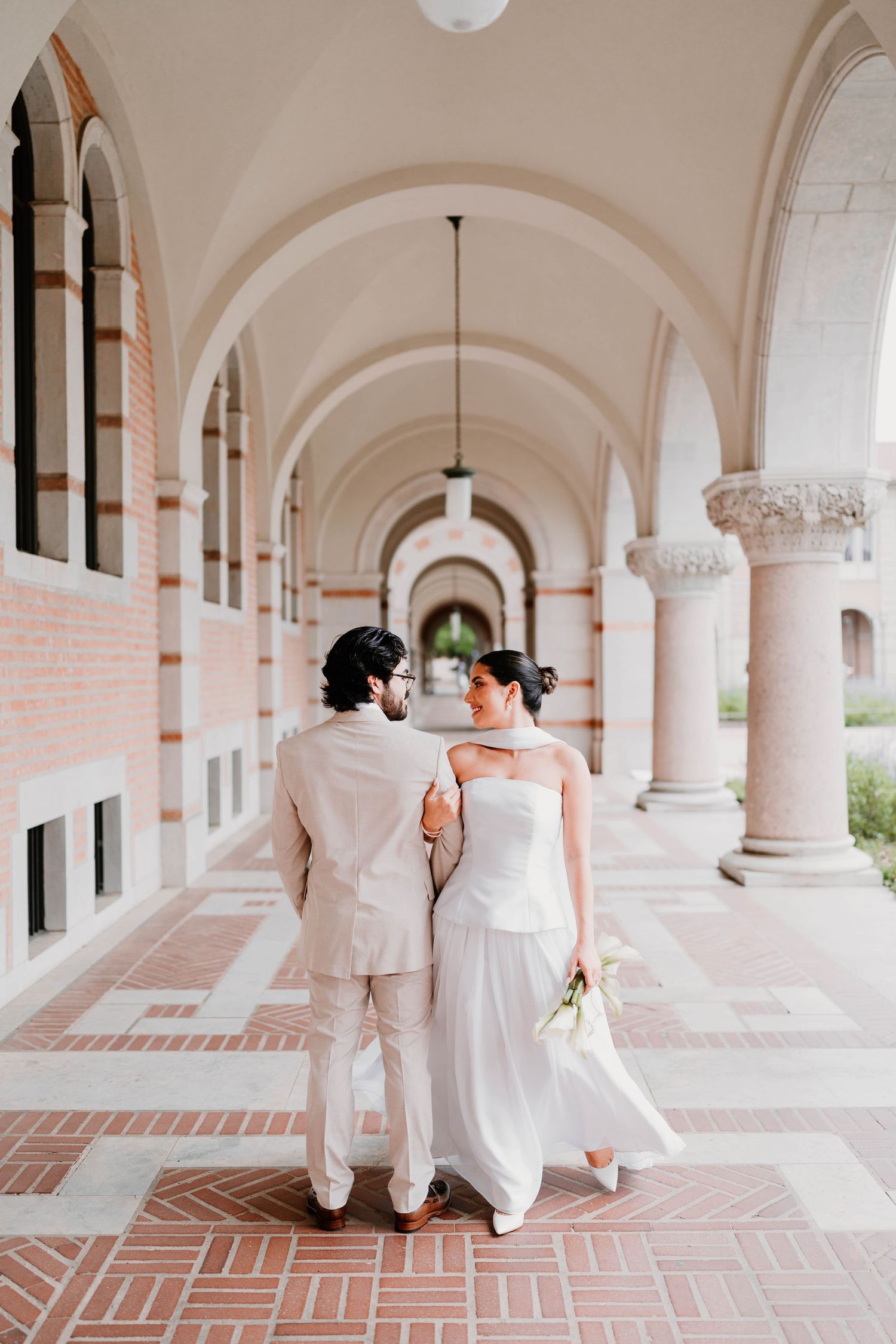 Bride and groom walk hand in hand through a beautiful columned corridor with brick detailing.