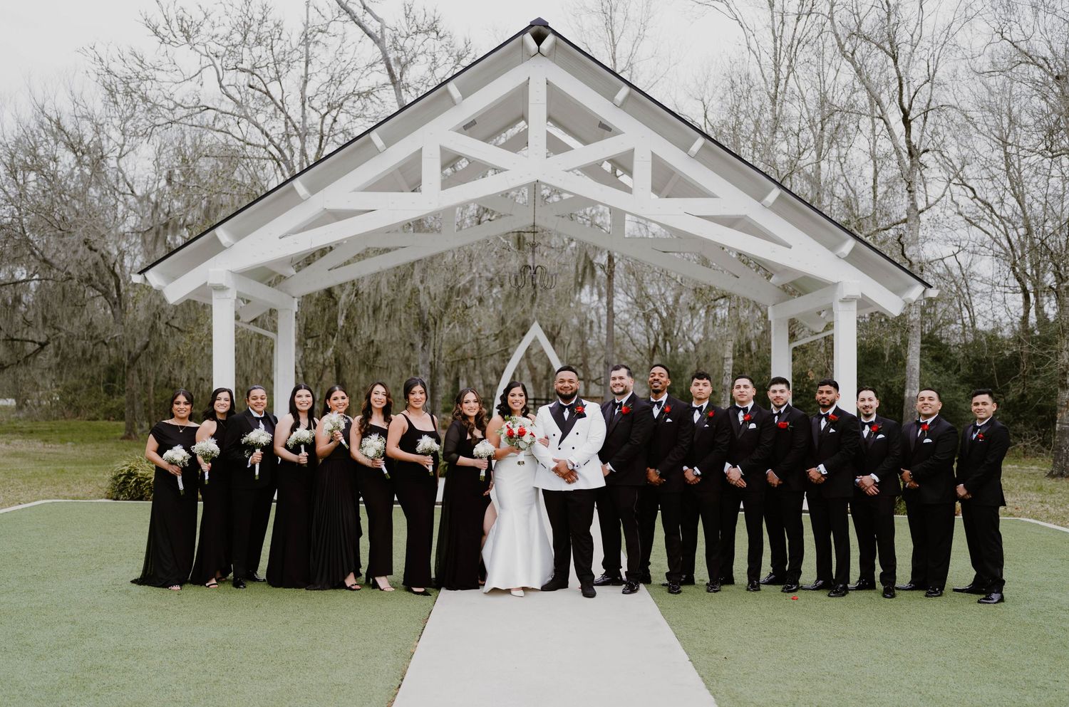 Large wedding party lined up for formal photos outside a white chapel structure.
