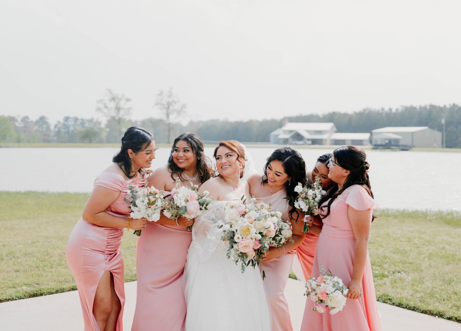 Bridesmaids in pink dresses pose together holding bouquets by a lake.