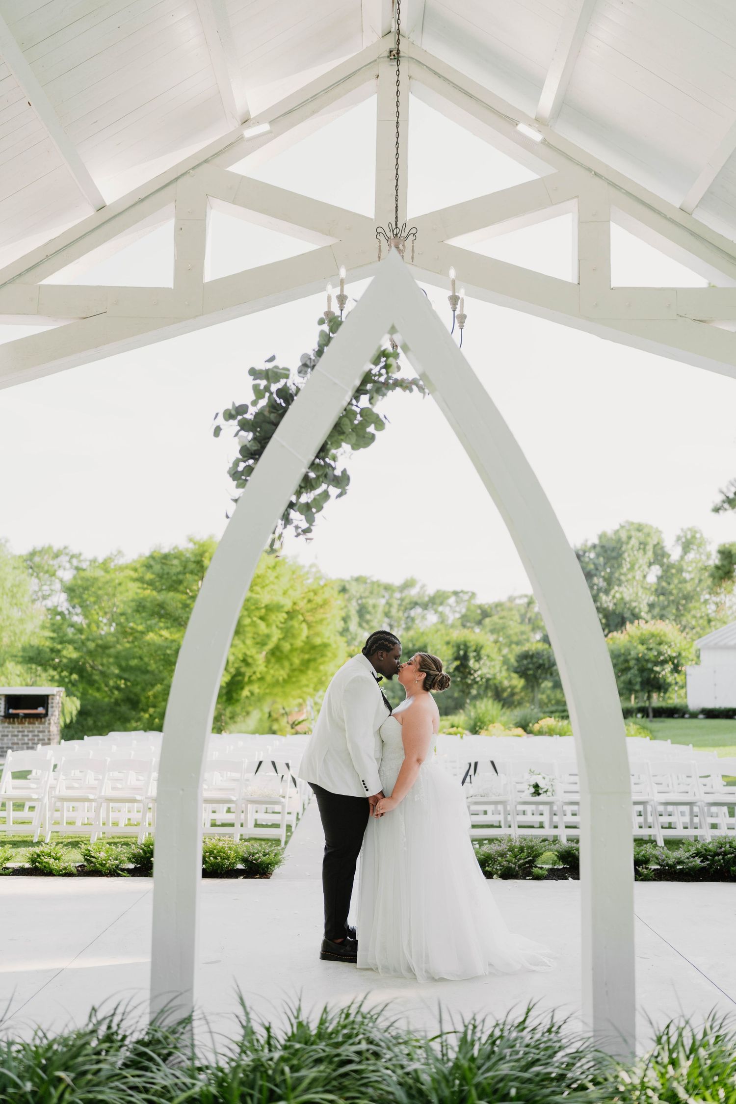 A couple shares a romantic moment under an elegant white wedding arch surrounded by lush greenery and white chairs.