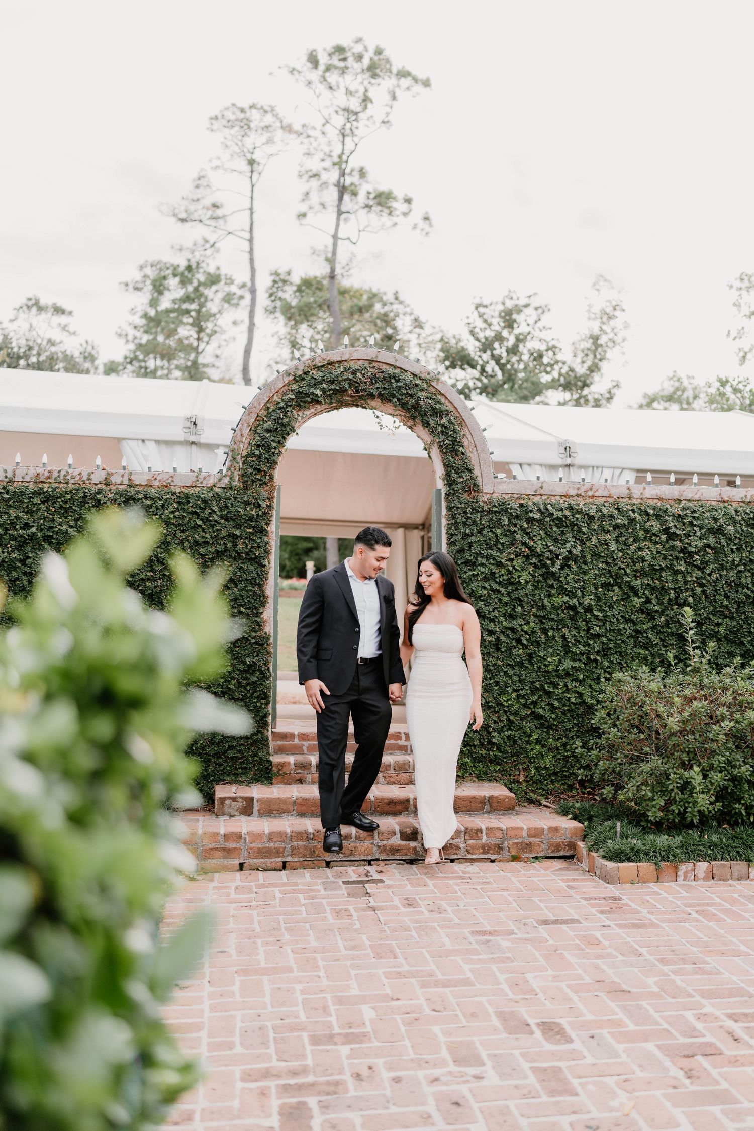 Couple walks along brick pathway next to manicured hedge wall in outdoor garden setting.