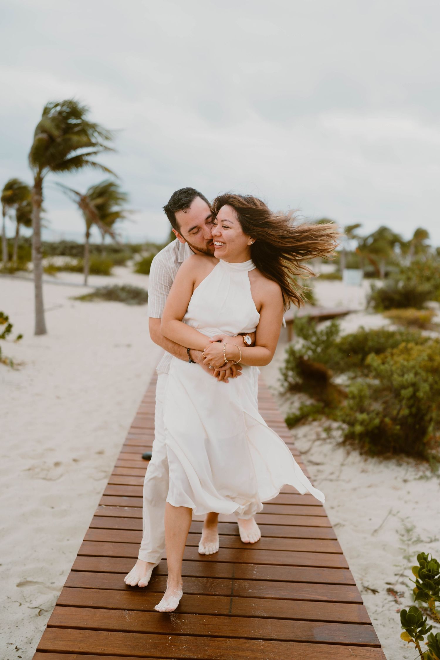 Romantic couple shares playful moments on wooden boardwalk with palm trees and beach in background.