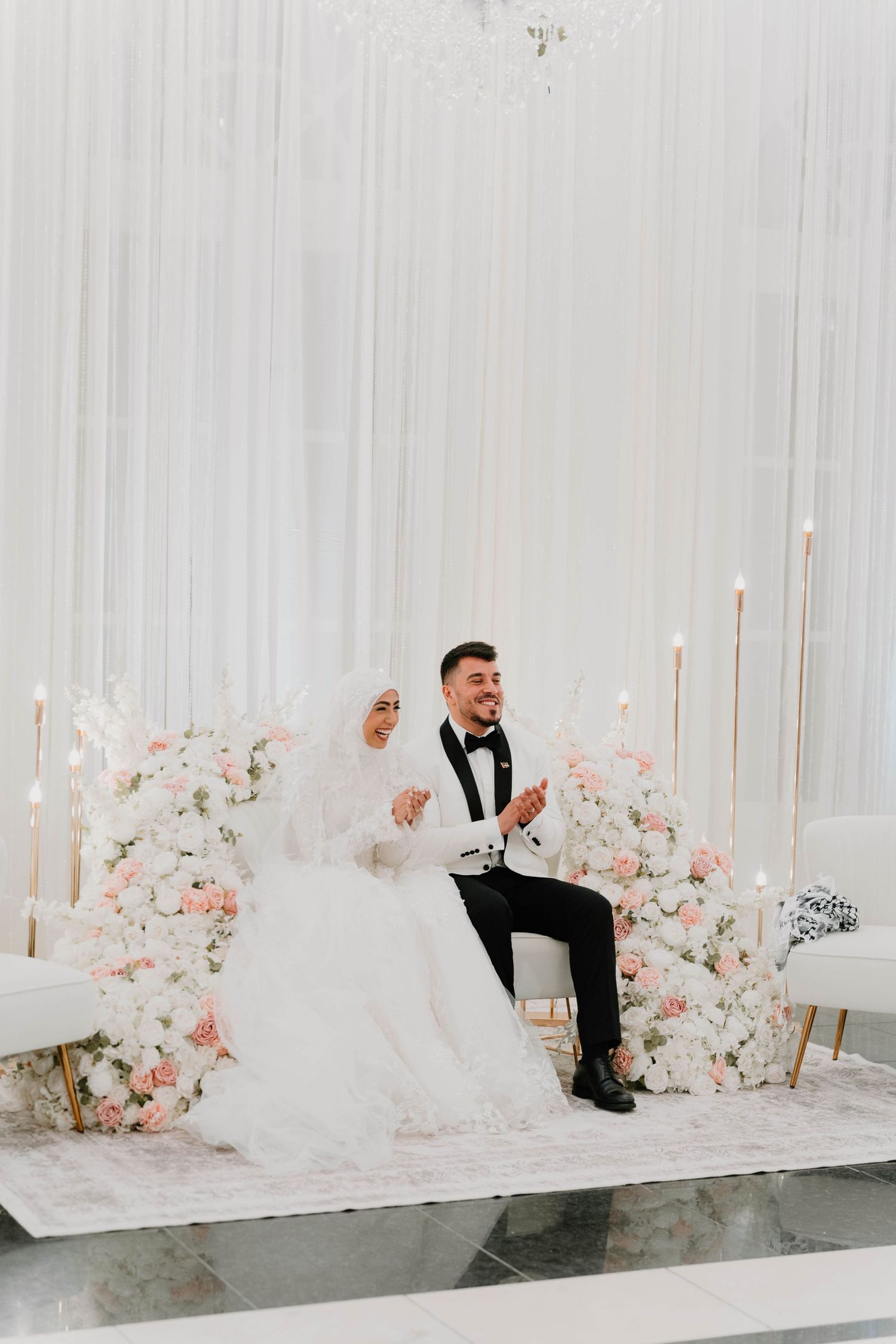 A couple sits on a decorated stage with pink floral arrangements and candles at their wedding reception.