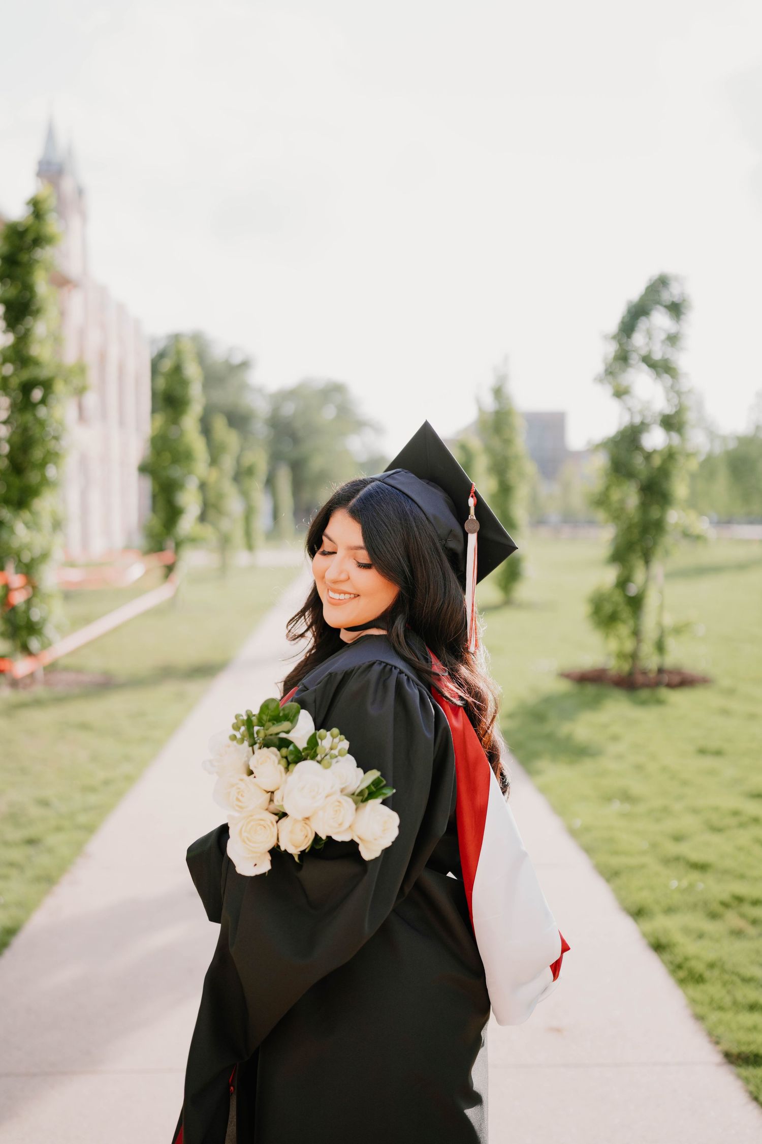 Graduate in black cap and gown poses with white flowers against a scenic hillside backdrop.