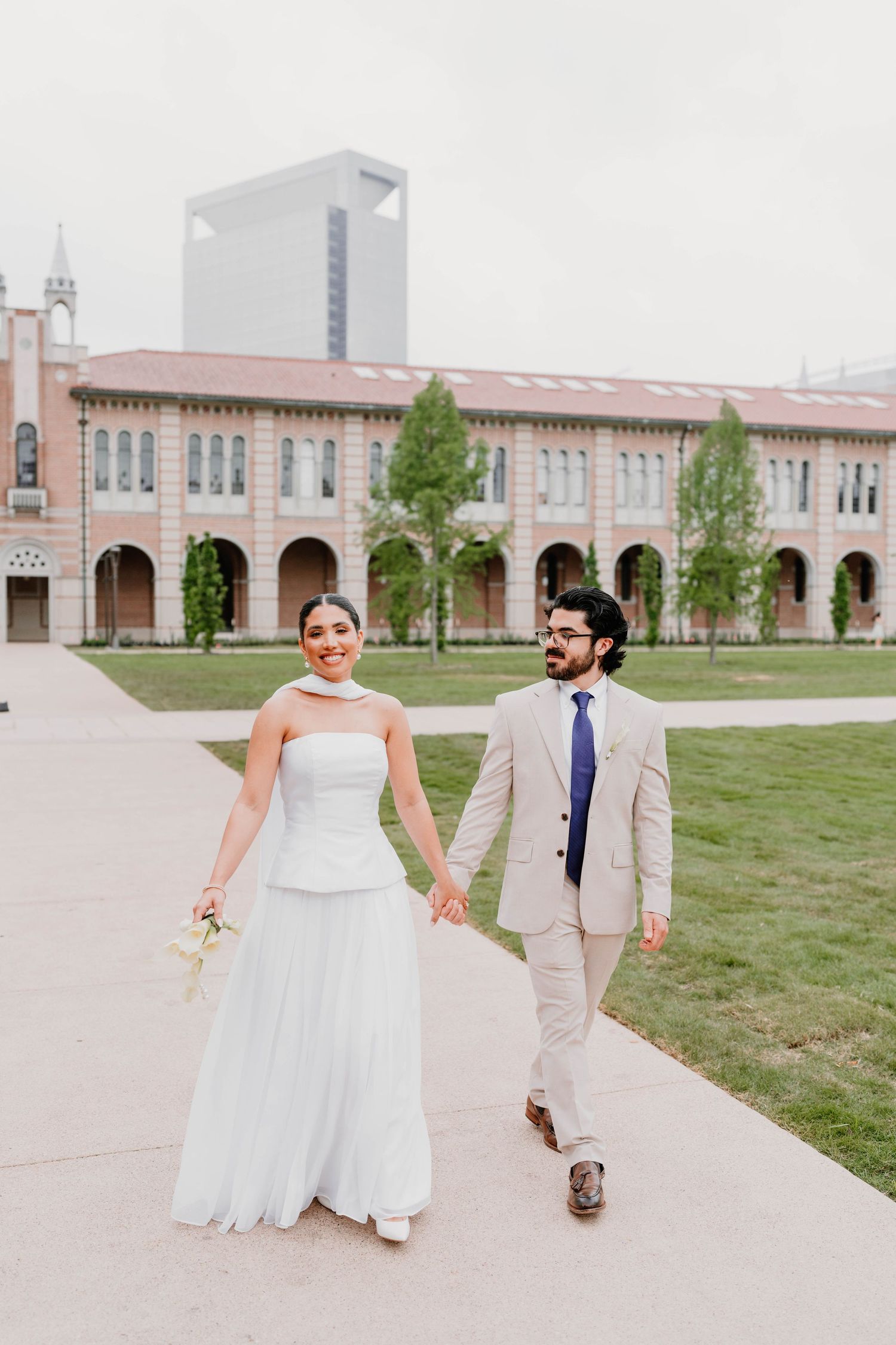 Wedding couple walks together on grass with historic building in background.