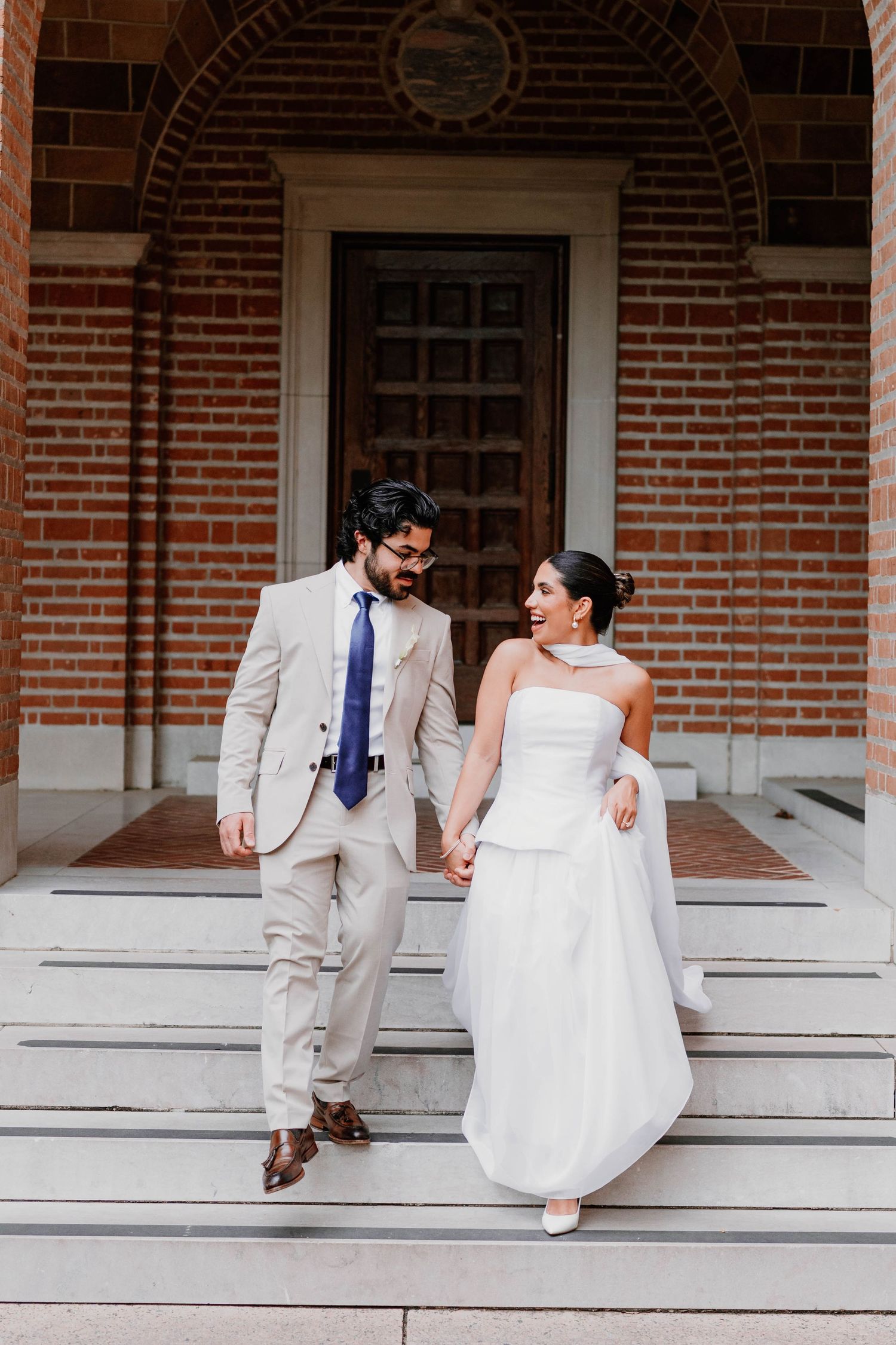 Wedding couple sits playfully on white steps of historic brick building.