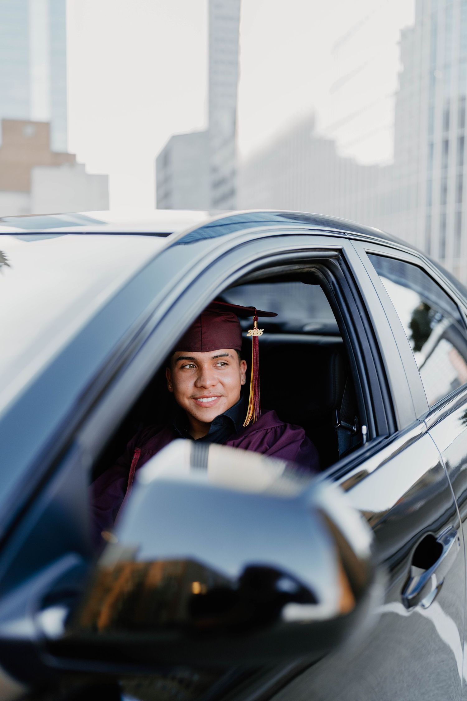 Graduate in maroon regalia sitting in car with city buildings in background.