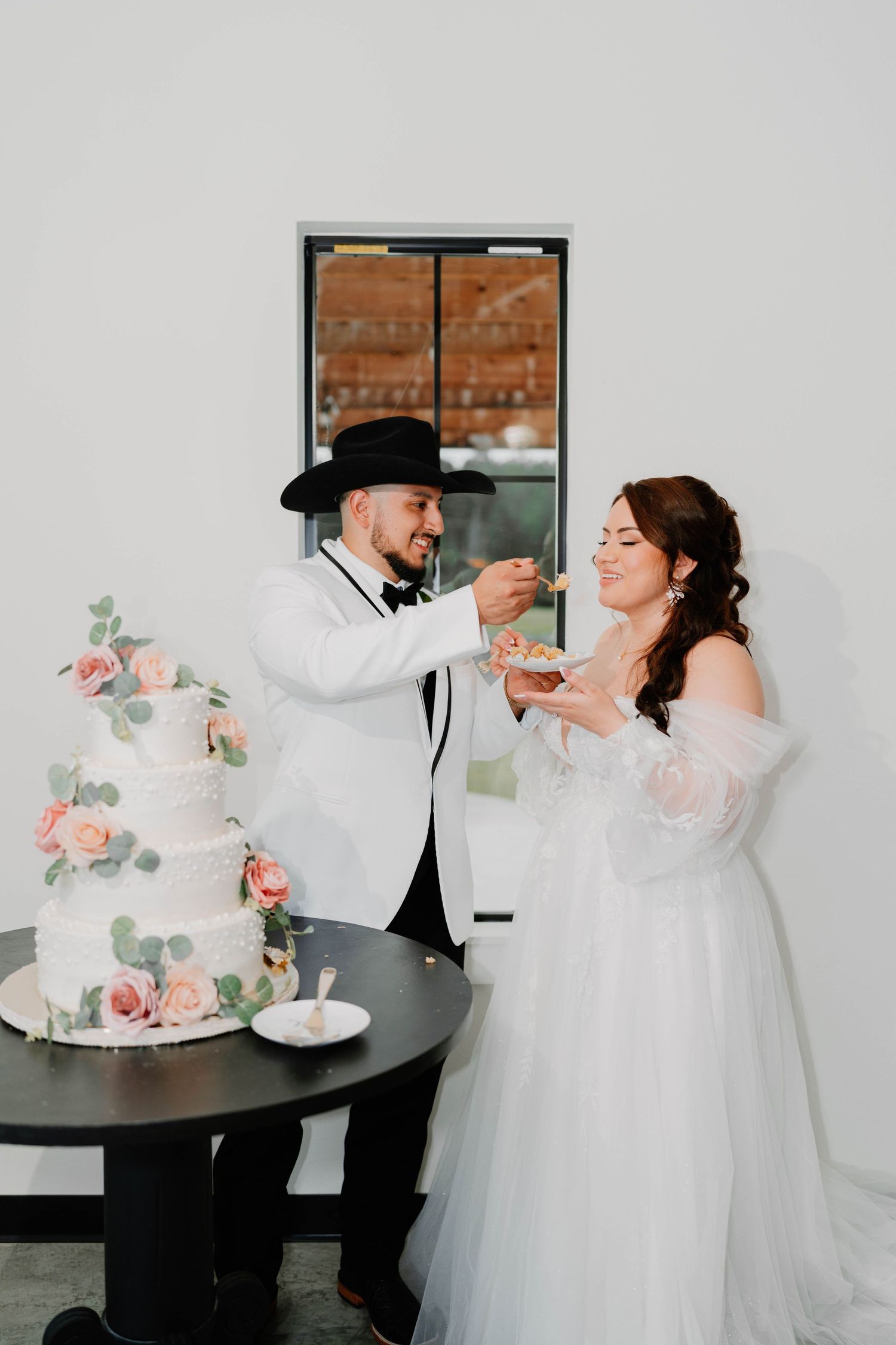 Newlyweds share cake at their wedding reception beside a floral decorated table.