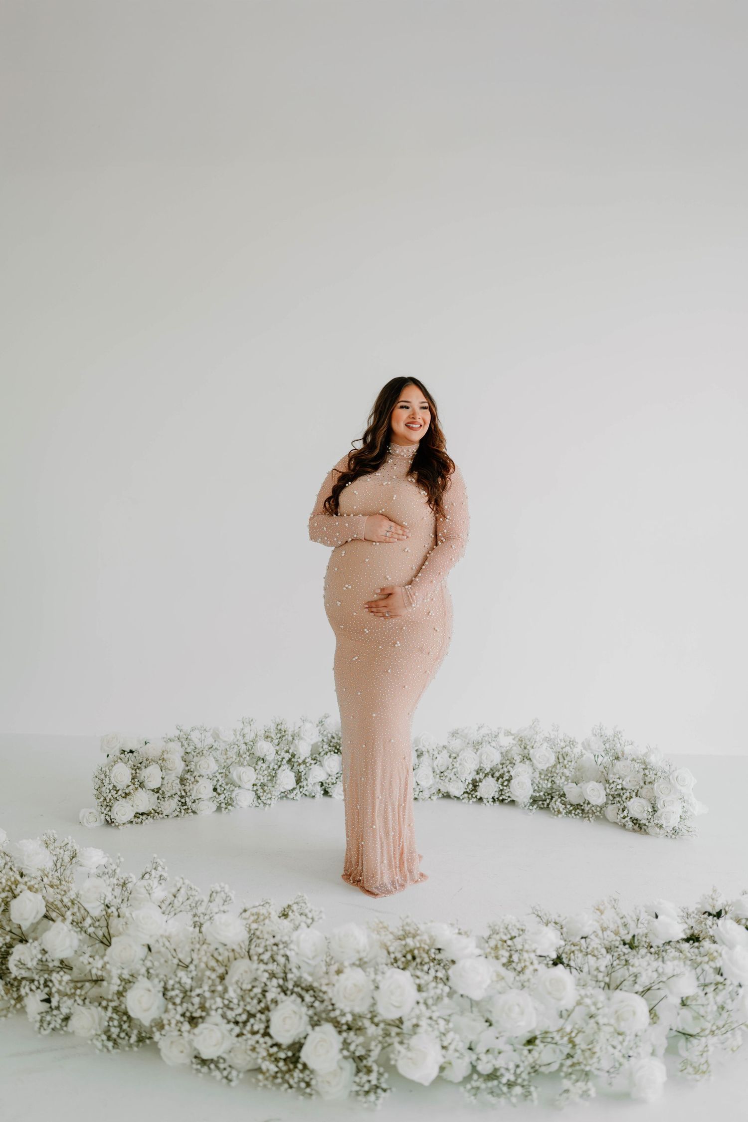 Maternity photo with white floral arrangements creating romantic setting against white backdrop.