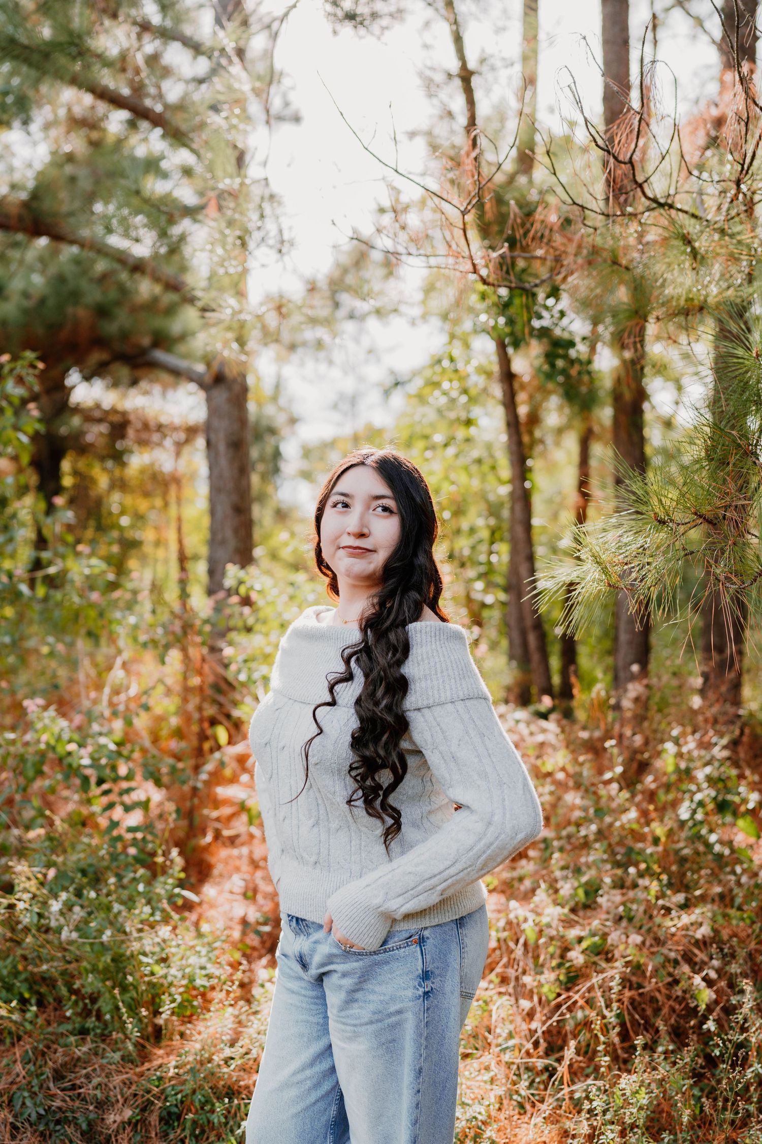 Young woman with long curly dark hair in a white sweater and jeans poses in a lush autumn forest setting.