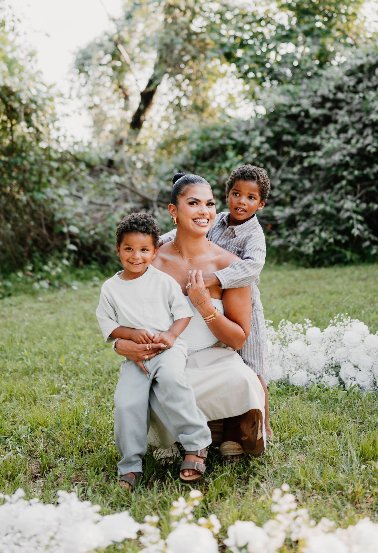 A tender family moment captured outdoors with white flowers blooming in the foreground on a sunny day.