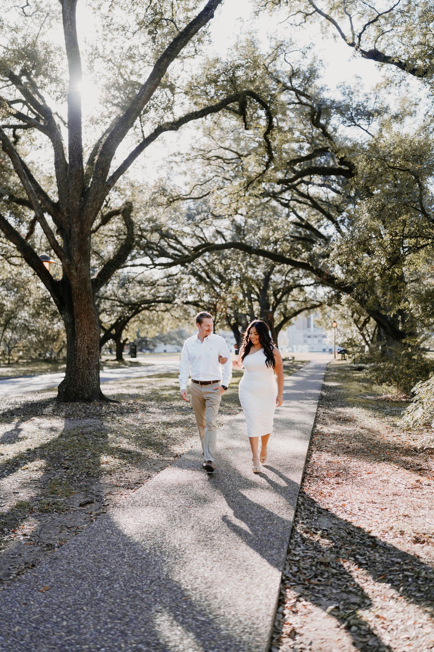 A couple walks hand in hand down a tree-lined path on a bright sunny afternoon.