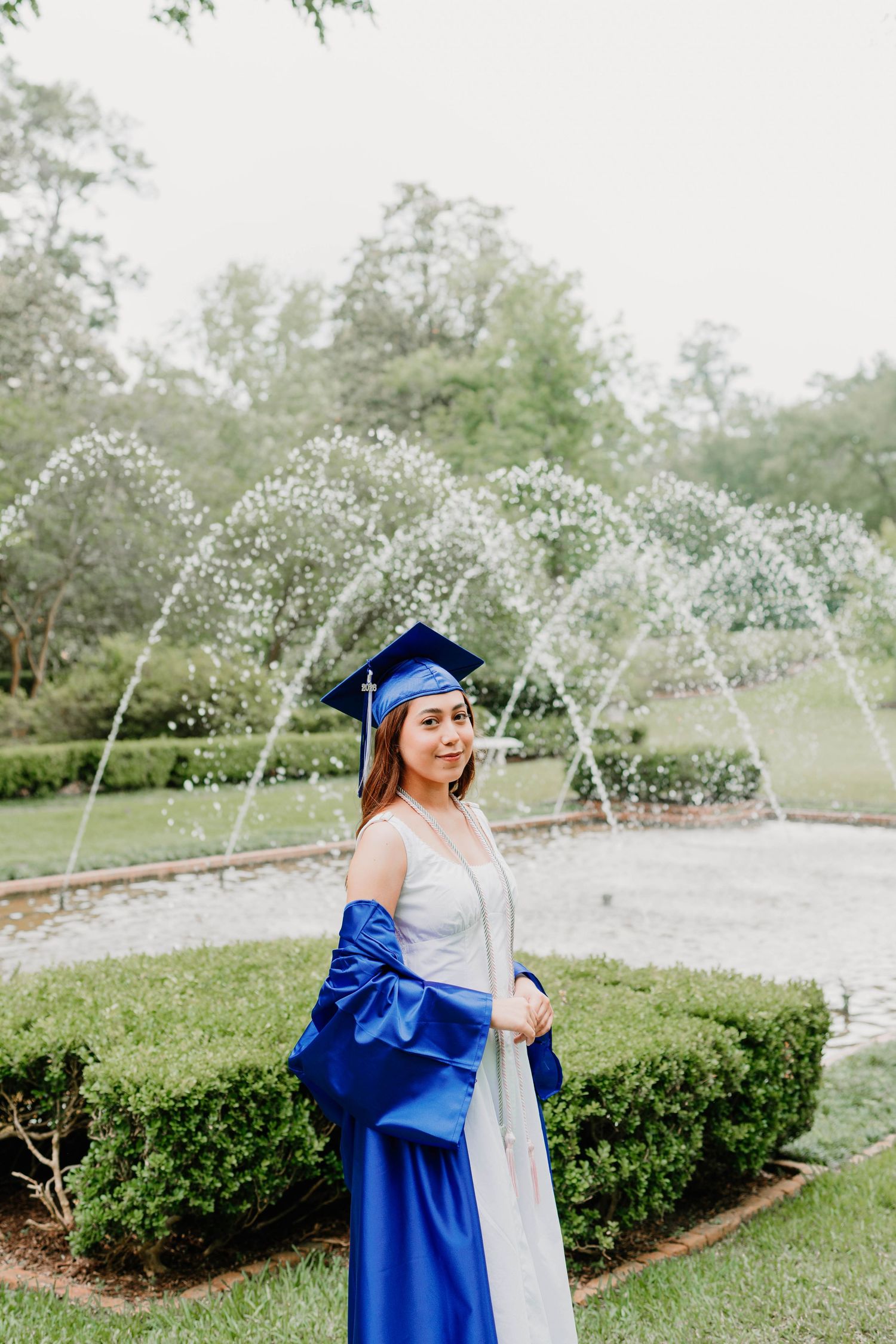 Young woman in blue graduation cap and gown smiling in front of decorative fountain surrounded by green hedges.