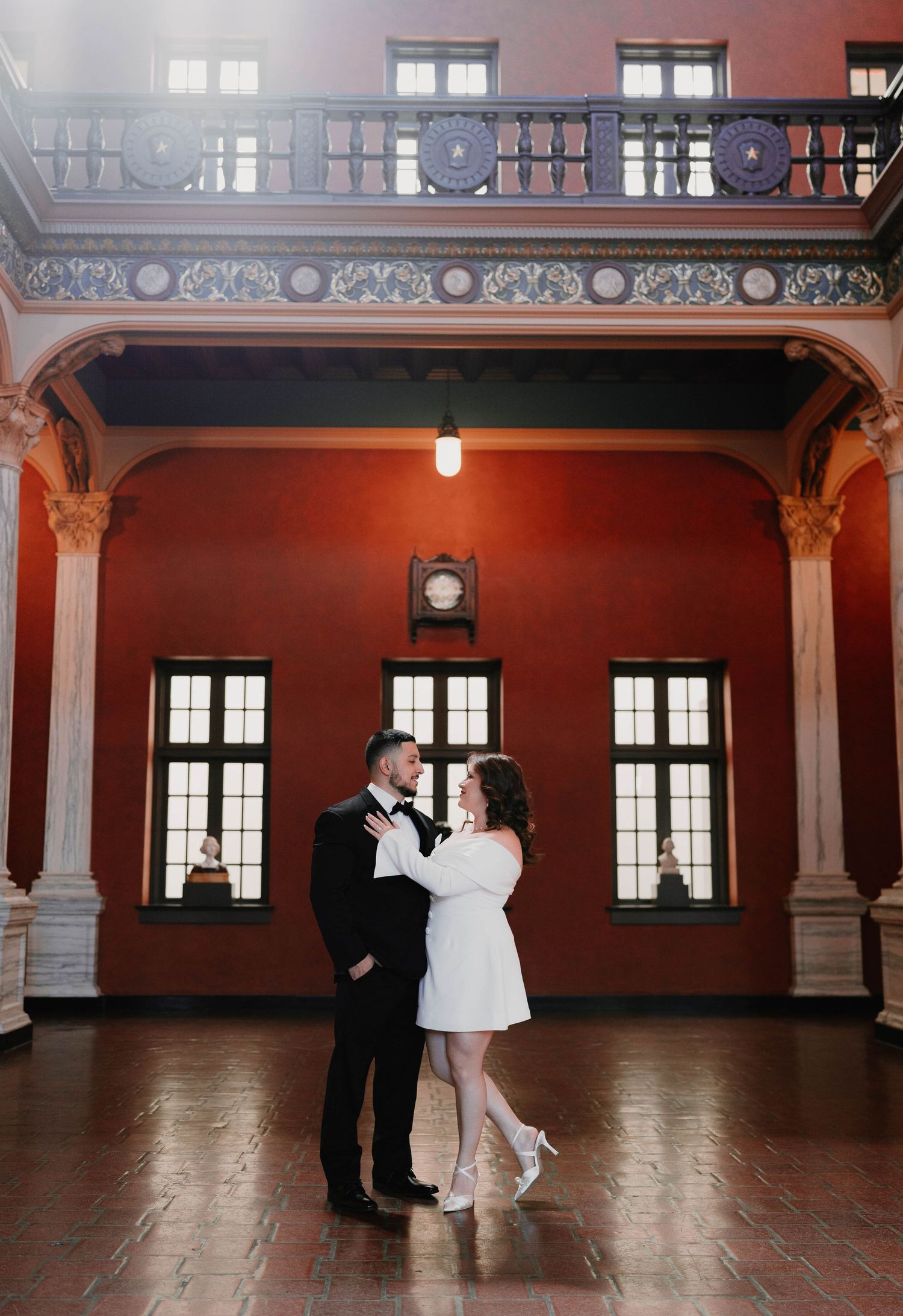 A couple dances together in a grand historic ballroom with ornate red walls and architectural details.