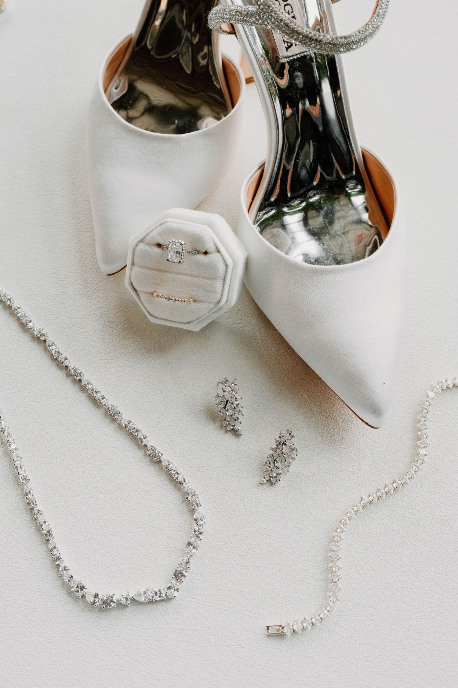 Wedding accessories including white heels, silver jewelry, and delicate bridal details arranged on white surface.