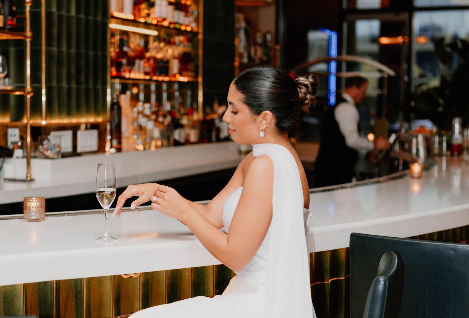 Bride in white dress sits at elegant bar counter with wine glass.