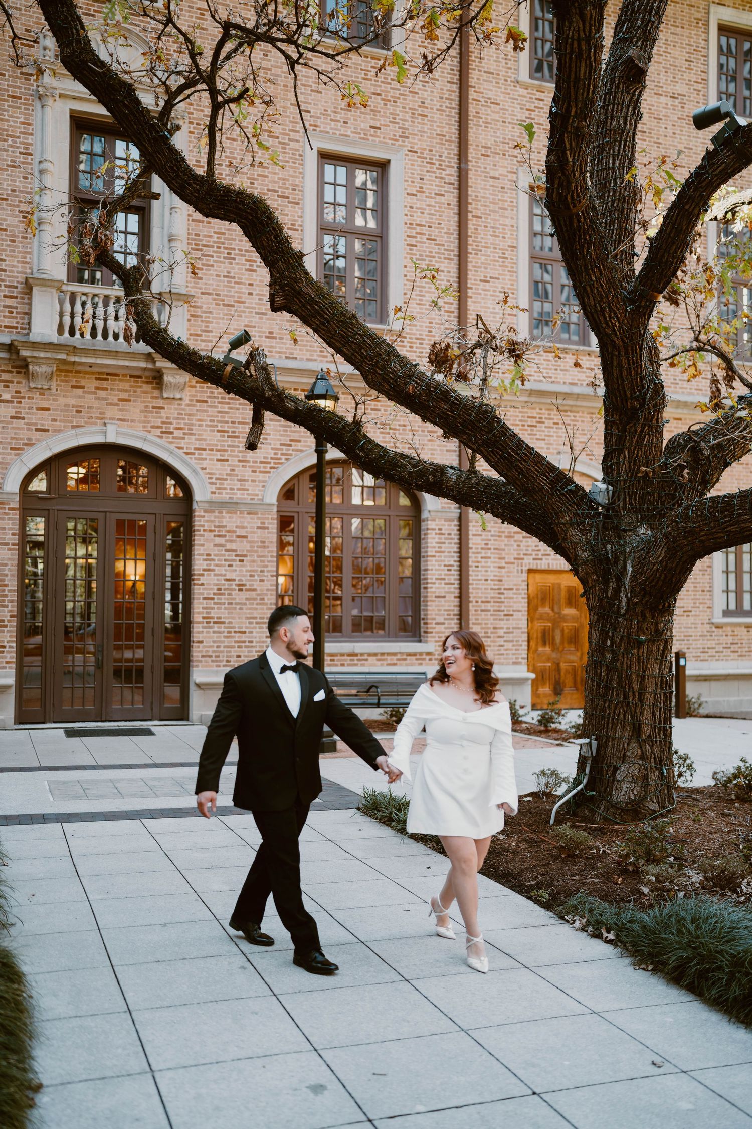 A couple walks hand in hand outside a brick building beneath a large twisted tree.