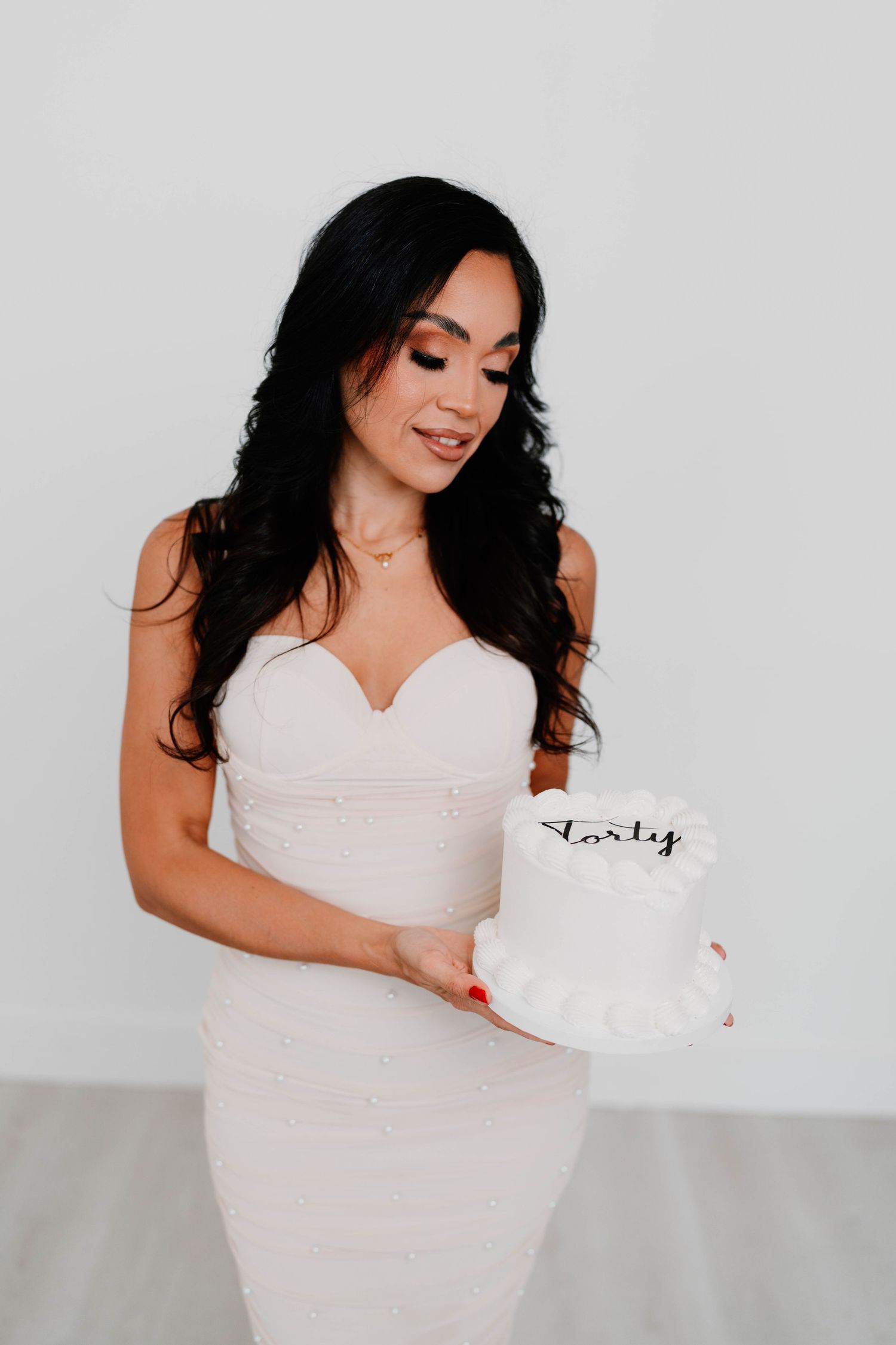Woman in white dress poses with a white cake against light background for elegant maternity photos.