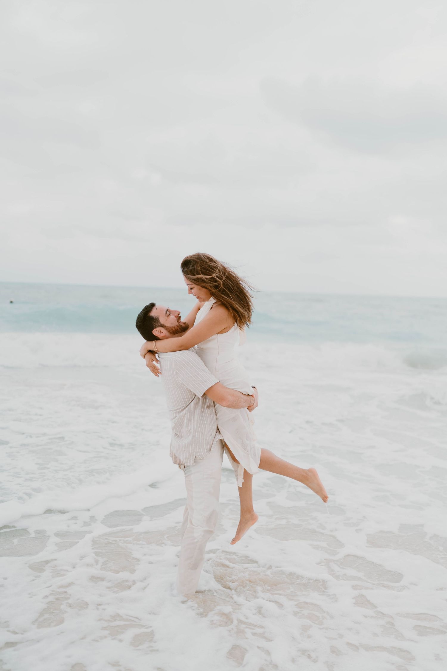 A romantic couple embracing on a beach with waves in the background during a cloudy day.