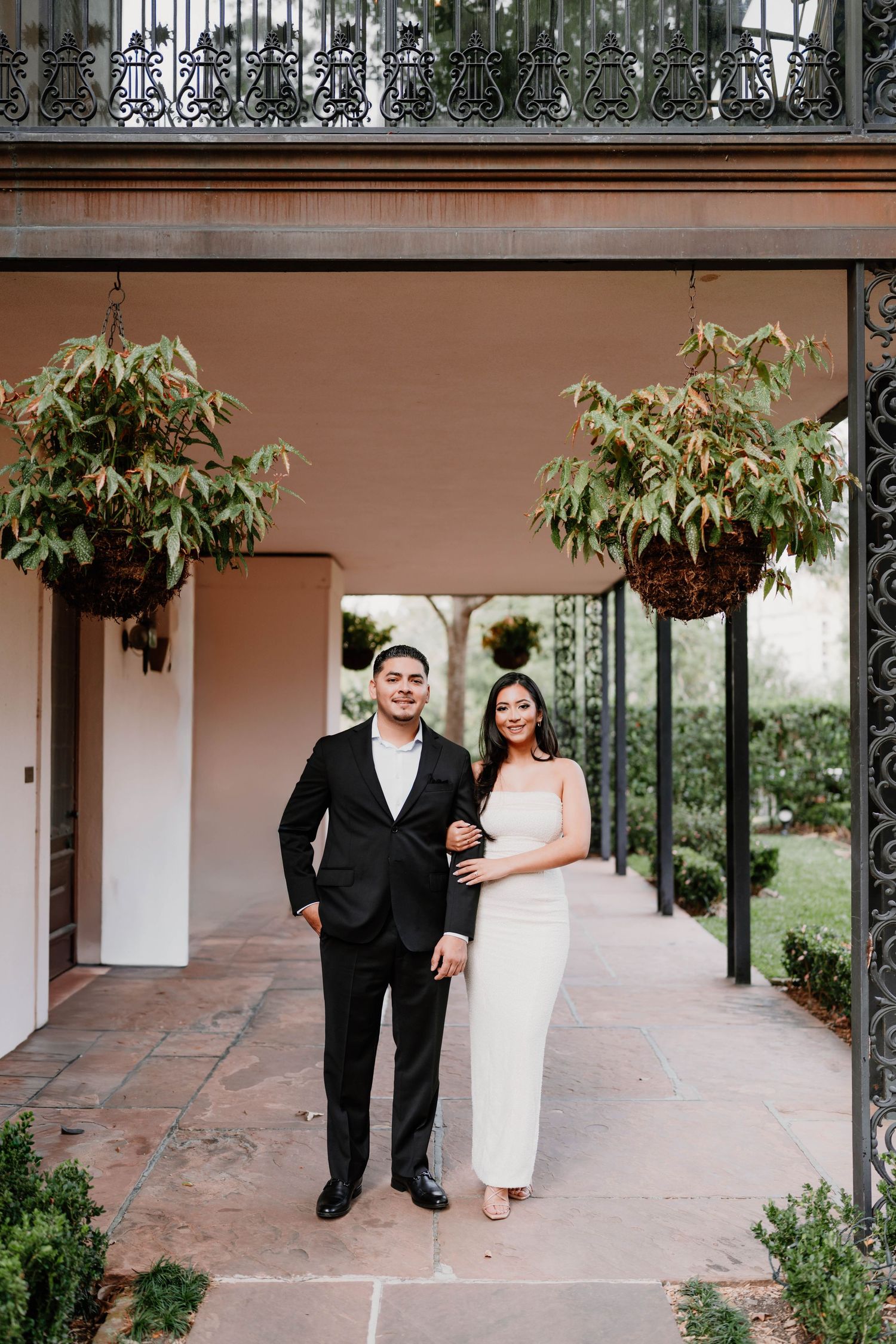 Formal couple walks under covered walkway with hanging plants and terracotta tiles.