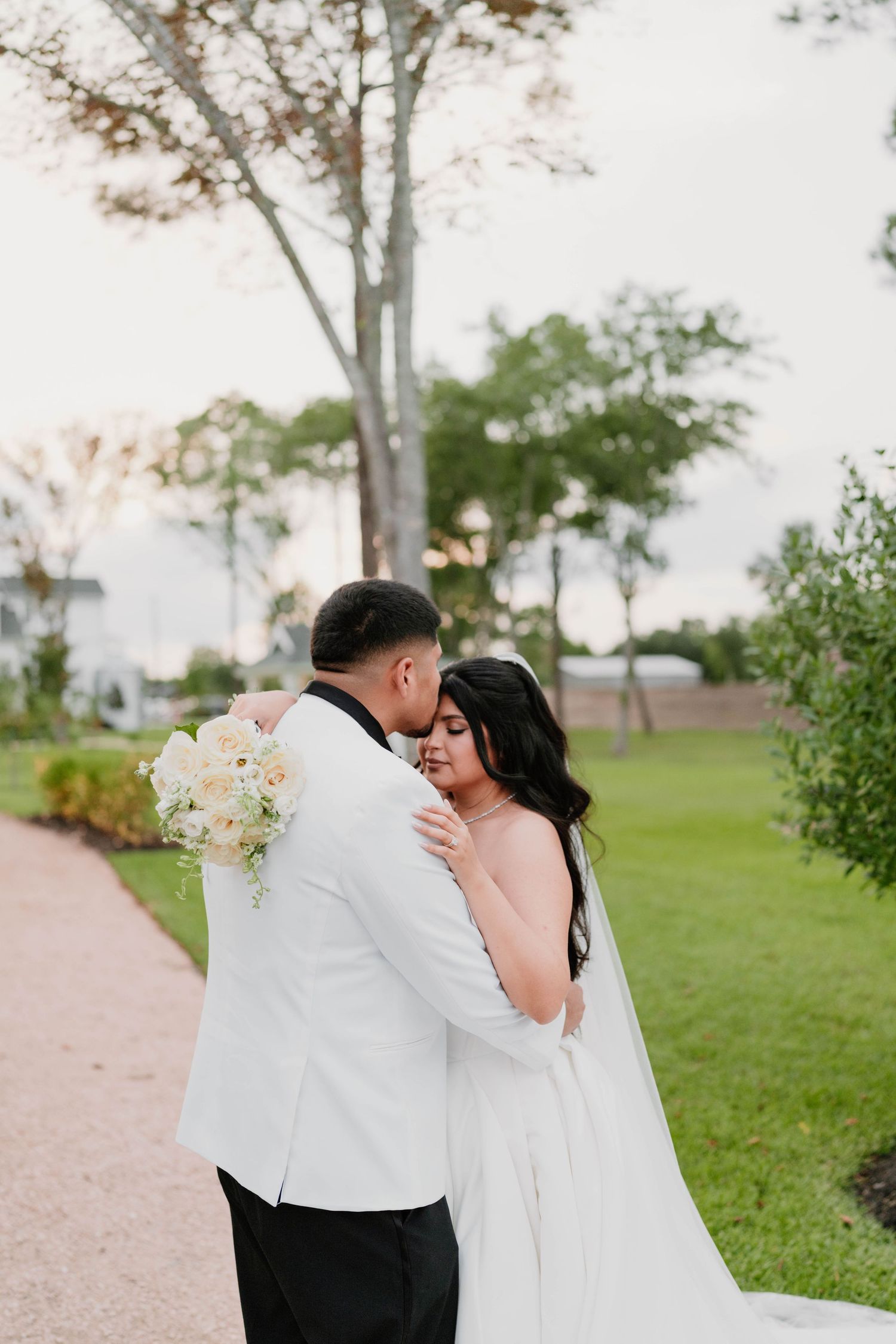 Wedding couple embracing on garden path with trees and greenery in background.