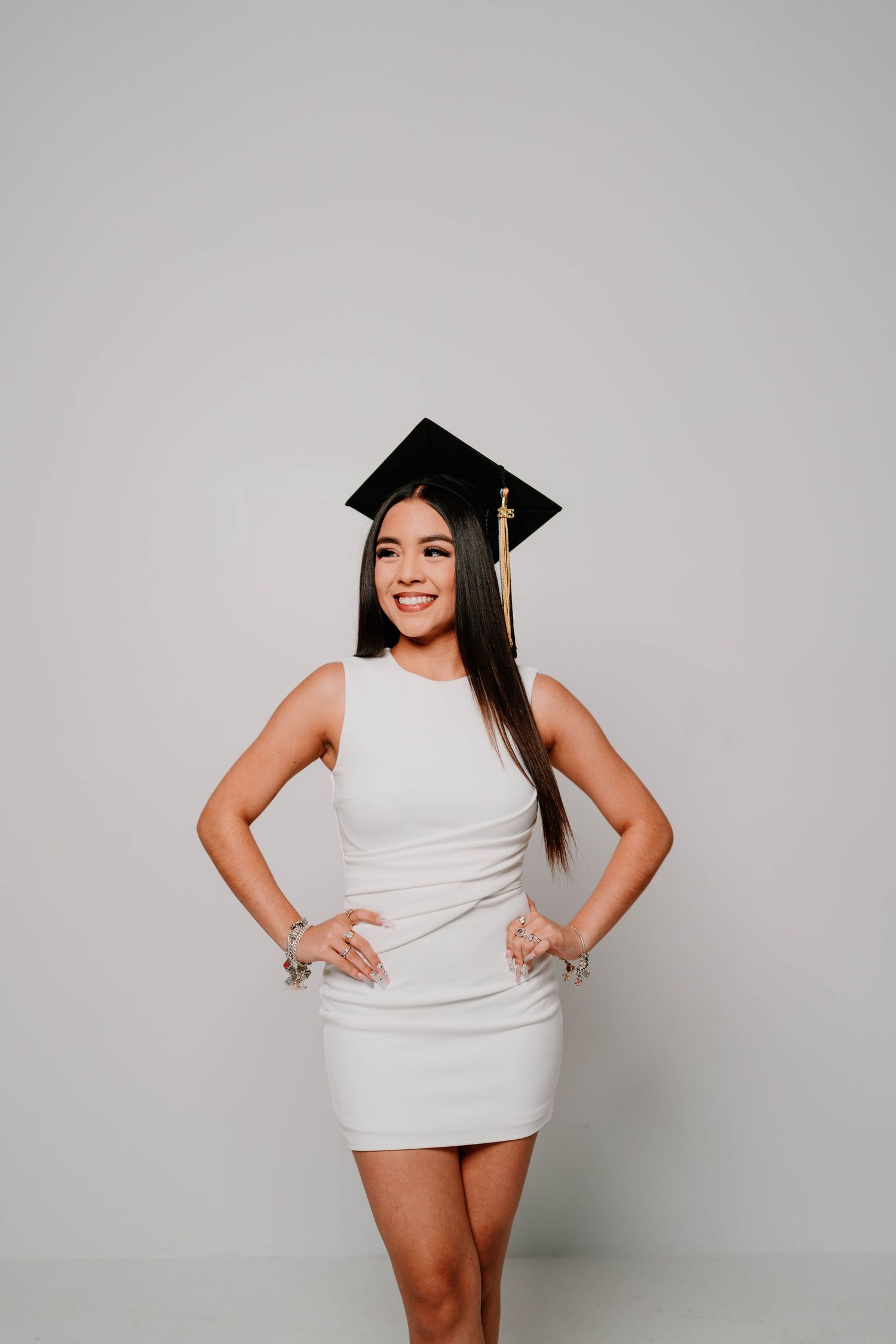 Celebratory graduation poses in white dress and cap against minimalist grey background.