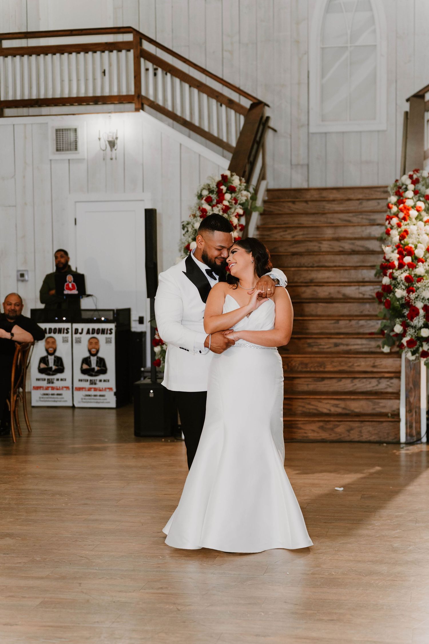 Wedding couple dances on wooden floor near decorated staircase in rustic white barn venue.