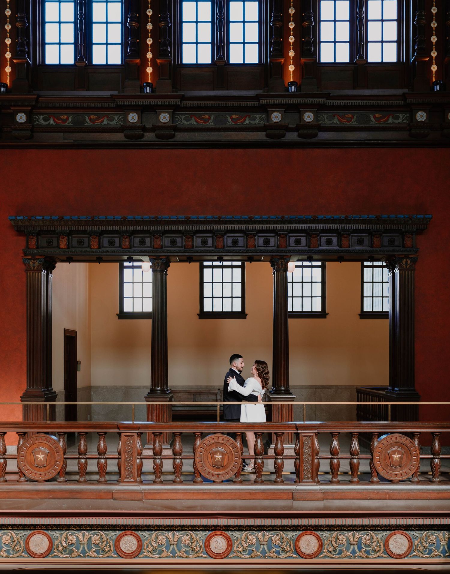 A couple embraces on a balcony in a historic building with dramatic red walls and ornate details.
