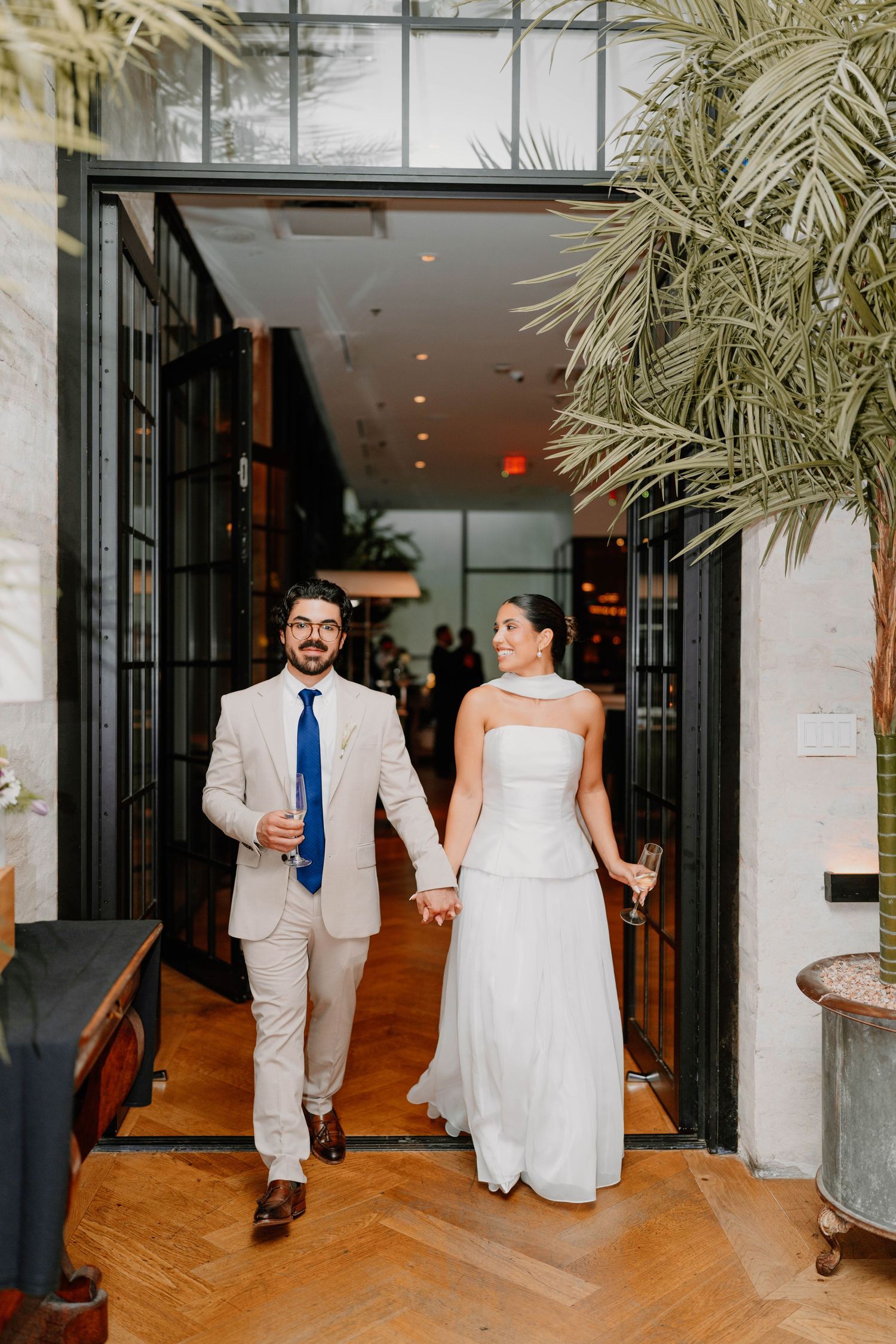 Newlyweds walk hand in hand through elegant venue with palm plants.