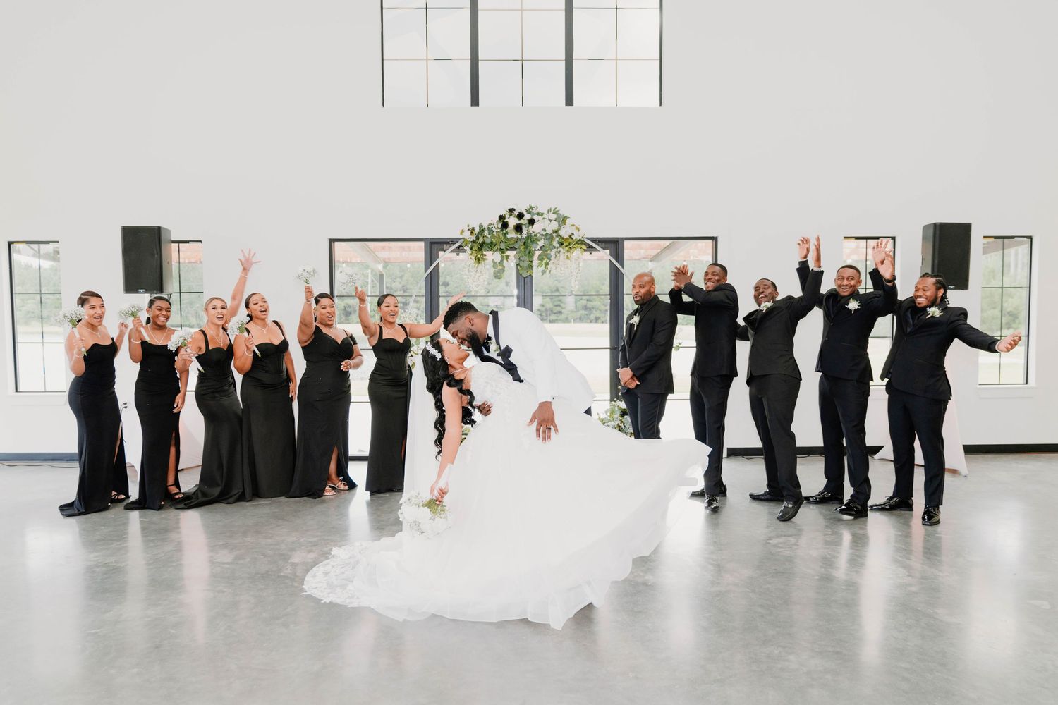 Wedding party celebrates as groom dips bride, bridesmaids in black gowns and groomsmen in tuxedos cheer in bright venue.