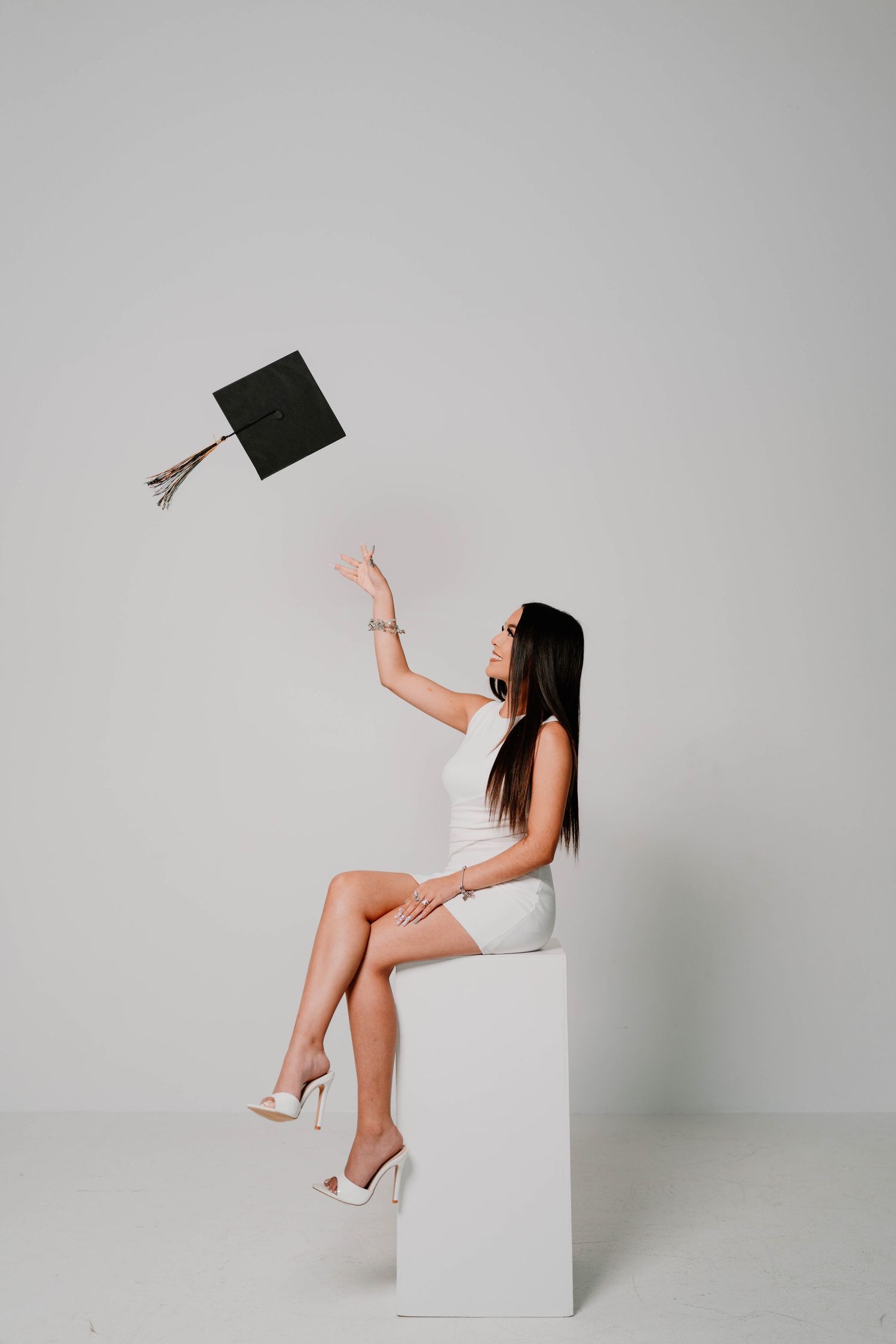 Playful graduation photo session with cap toss poses on white cube against grey background.