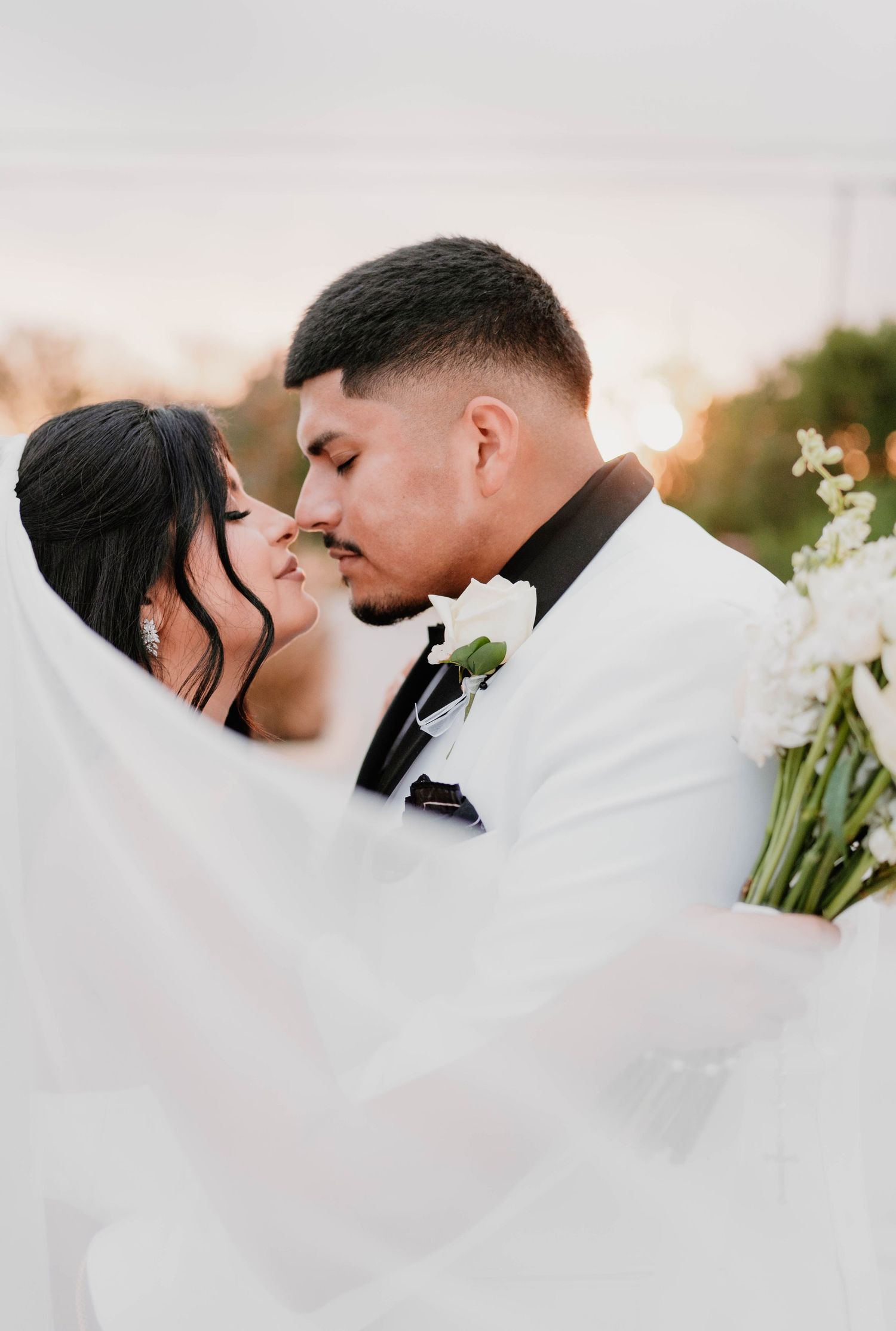 Intimate wedding portrait with bride's veil flowing in breeze during sunset.