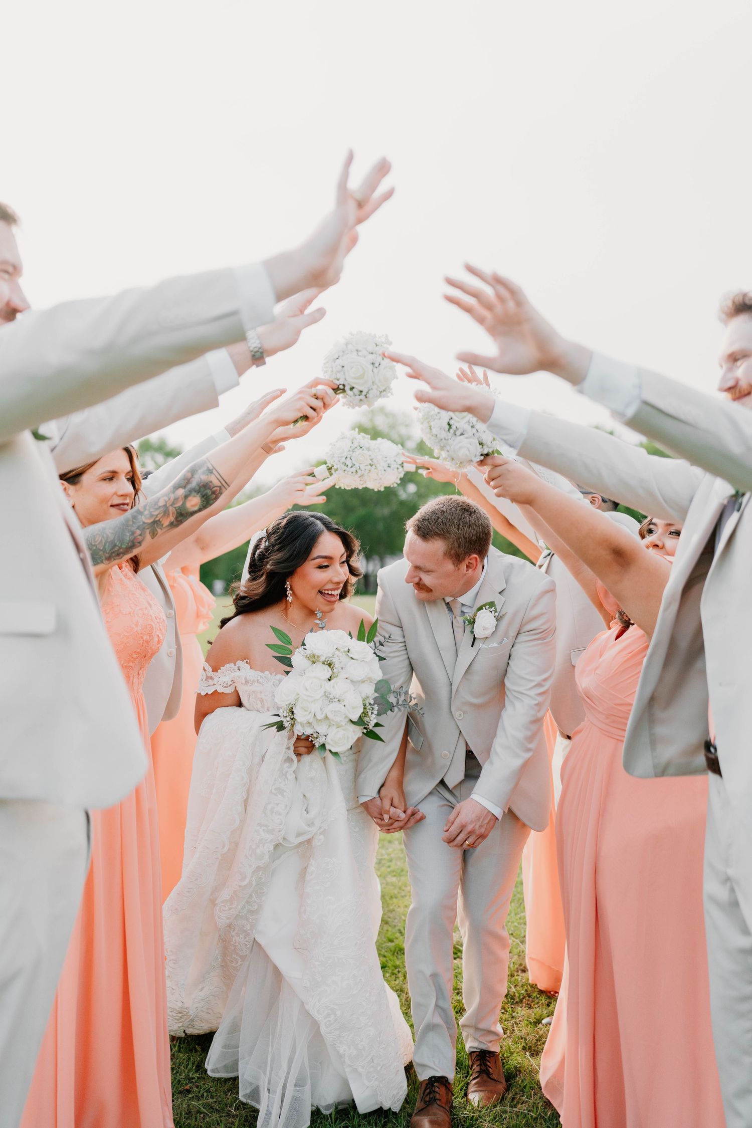 Wedding party in peach dresses and gray suits forms celebratory arch tunnel outdoors.