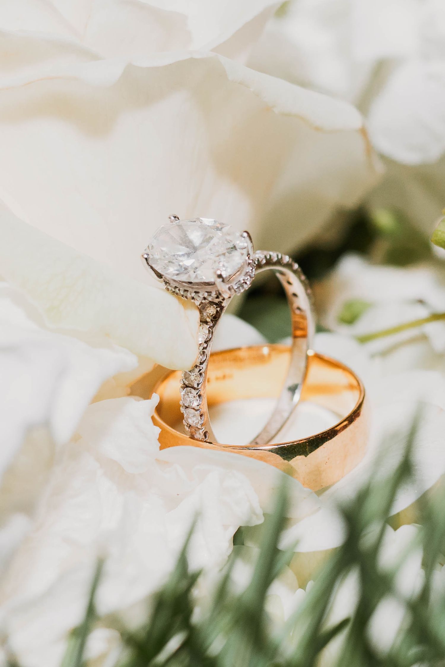 A sparkling diamond engagement ring rests delicately among white flower petals and green foliage.