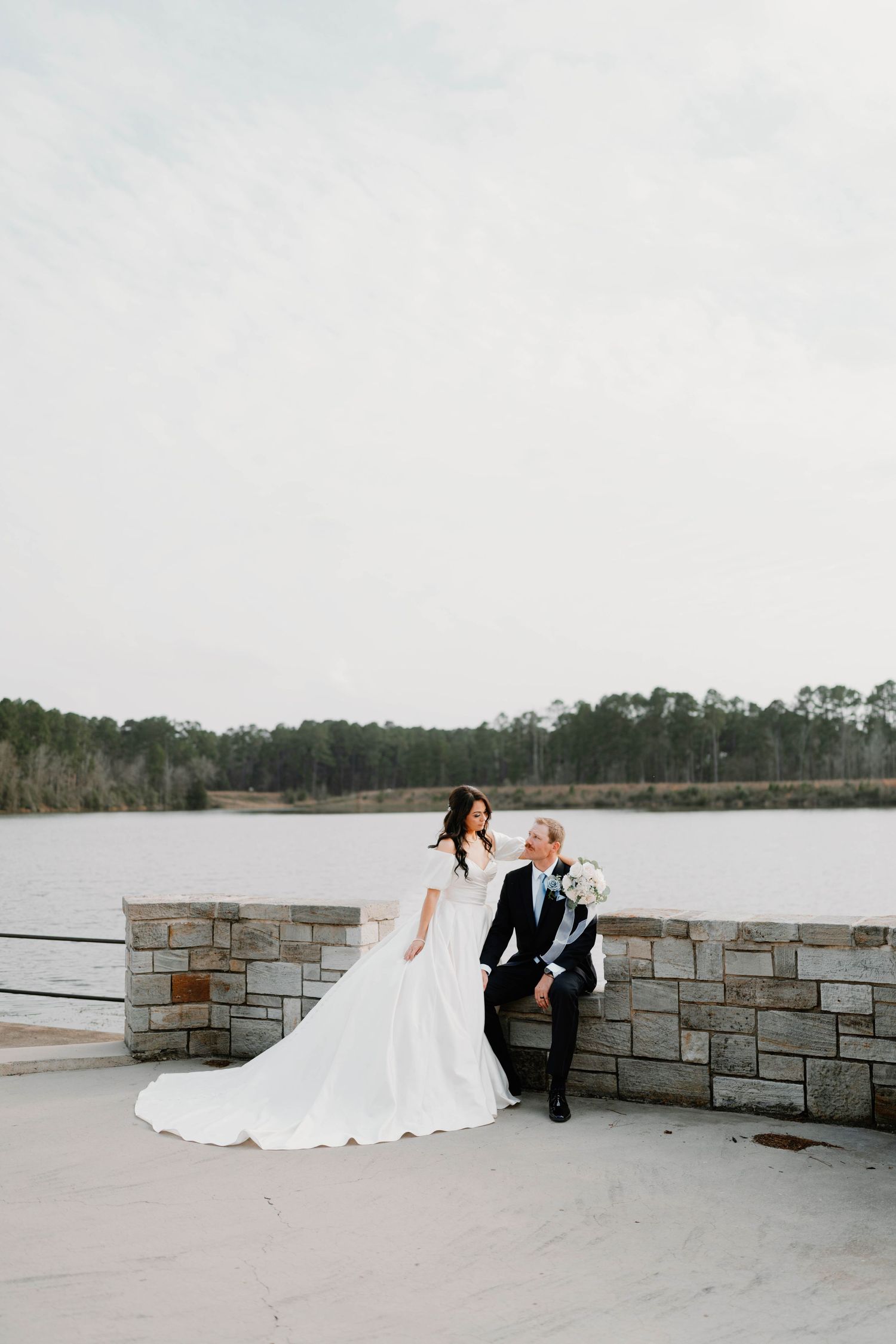Newlyweds share intimate moment on stone terrace with lake view backdrop.