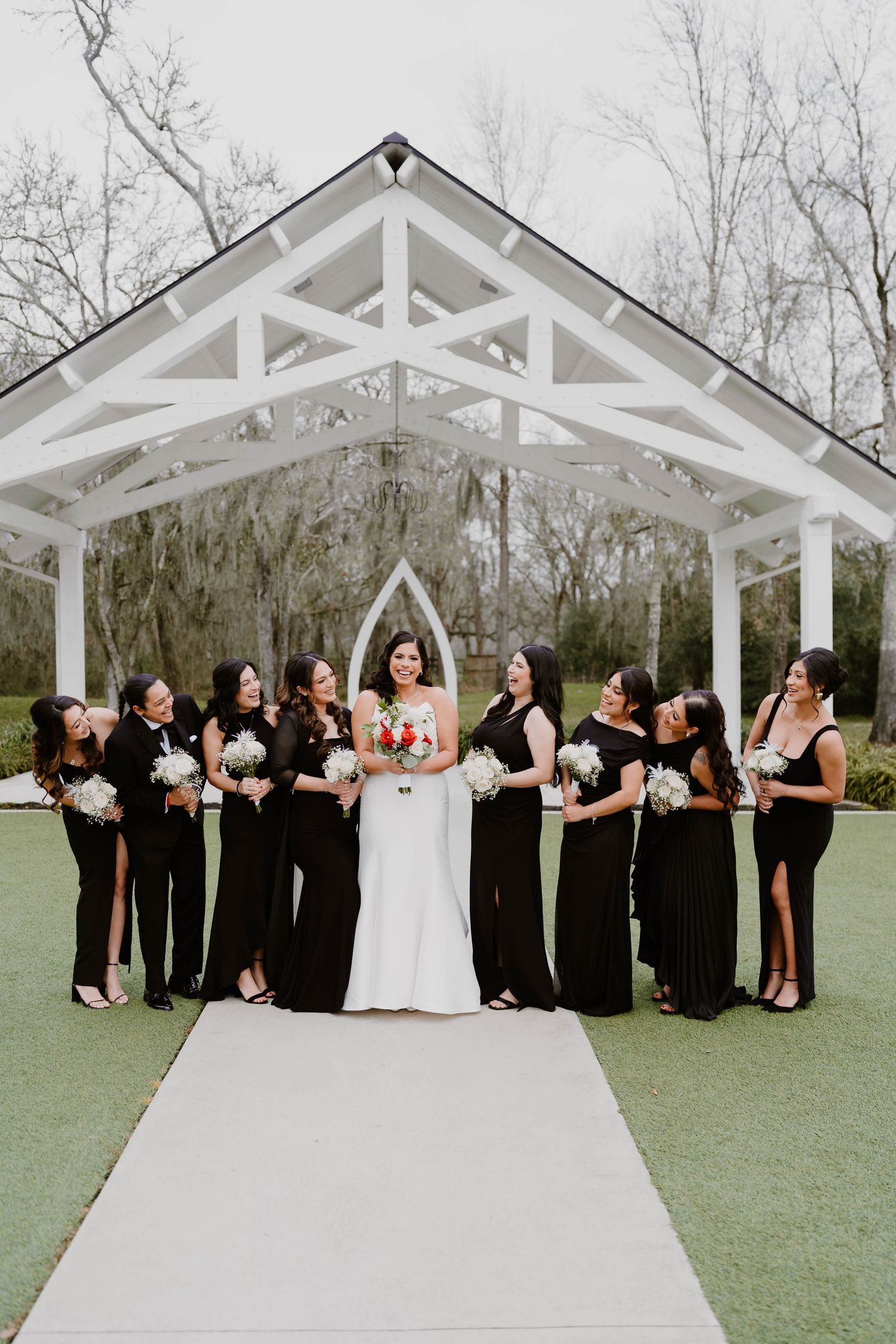 Wedding party in black attire poses in front of a white chapel structure outdoors.
