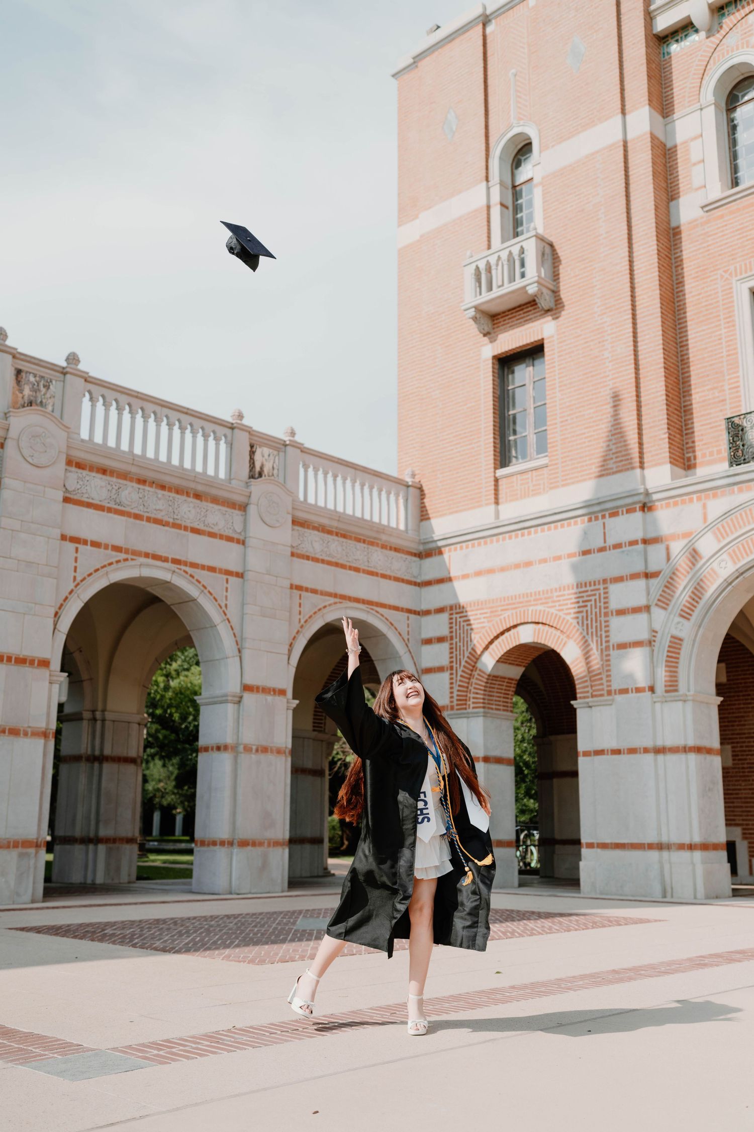 A graduate in cap and gown joyfully tosses her mortarboard in the air outside a classic university building.