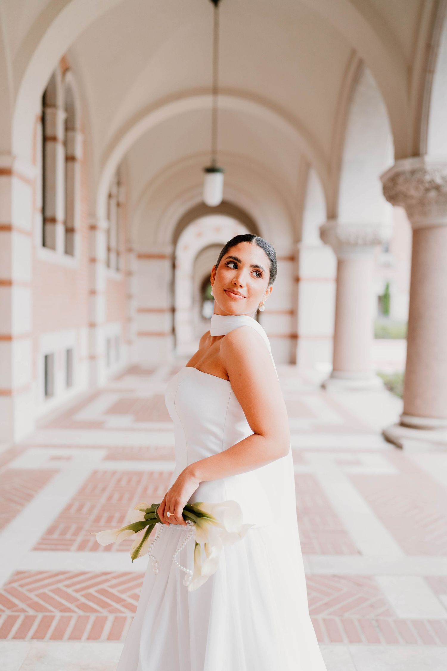 Bride in white dress poses gracefully in an elegant arched hallway.