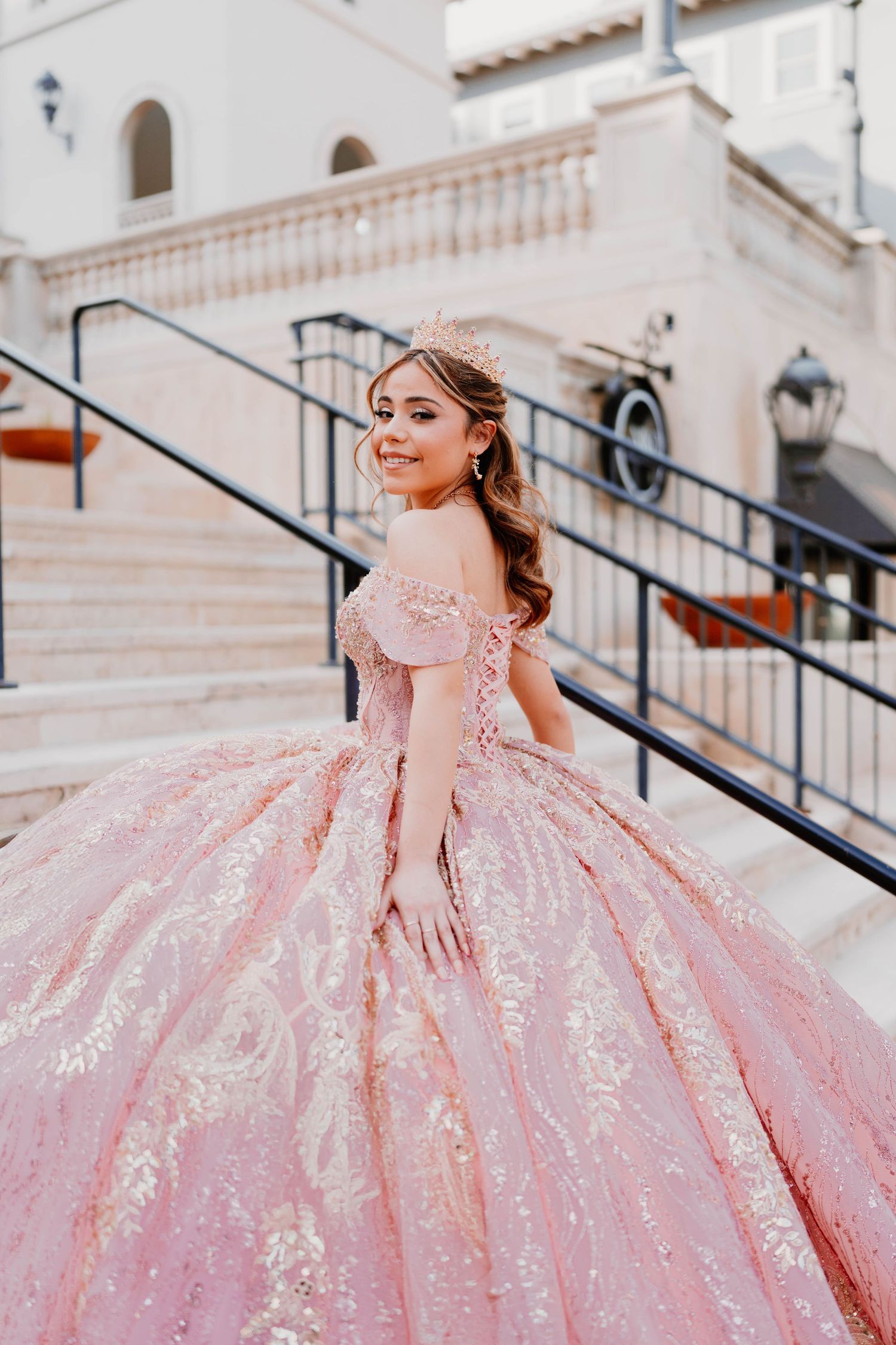Sparkling pink princess gown flows down elegant white stone steps during glamorous photoshoot.